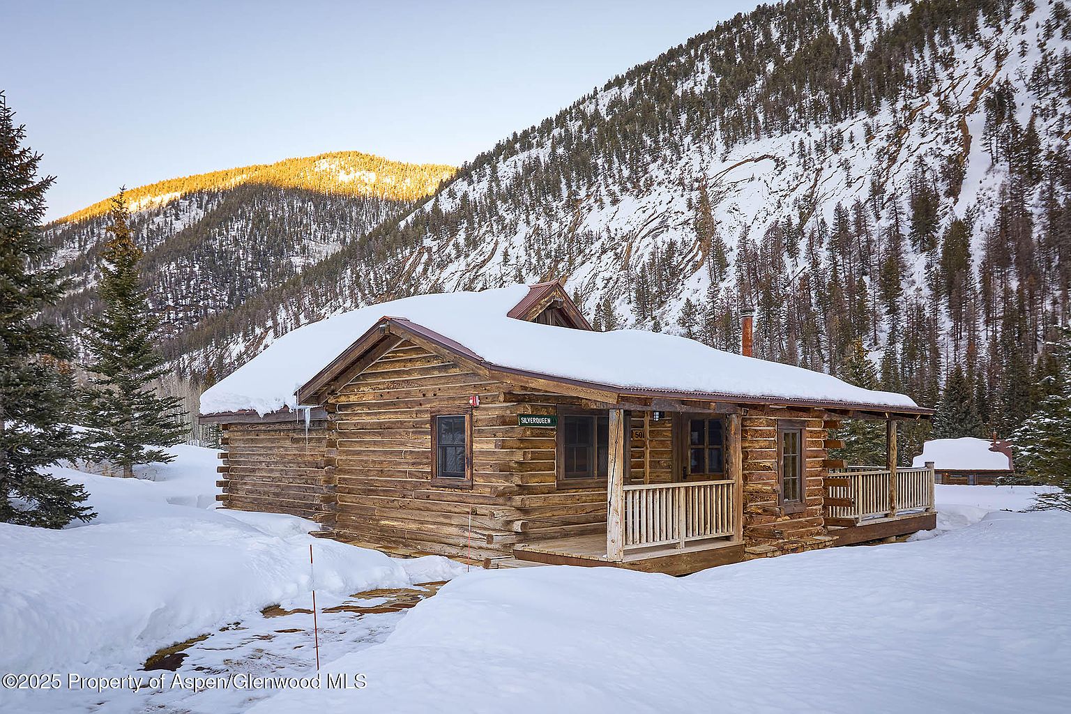 The image showcases a charming log cabin nestled in a snowy, mountainous landscape. The cabin features a snow-covered roof, a small porch, and visible log construction, exuding a rustic and cozy atmosphere. The surrounding snow-covered terrain and towering mountains create a picturesque and serene setting, ideal for a secluded retreat.