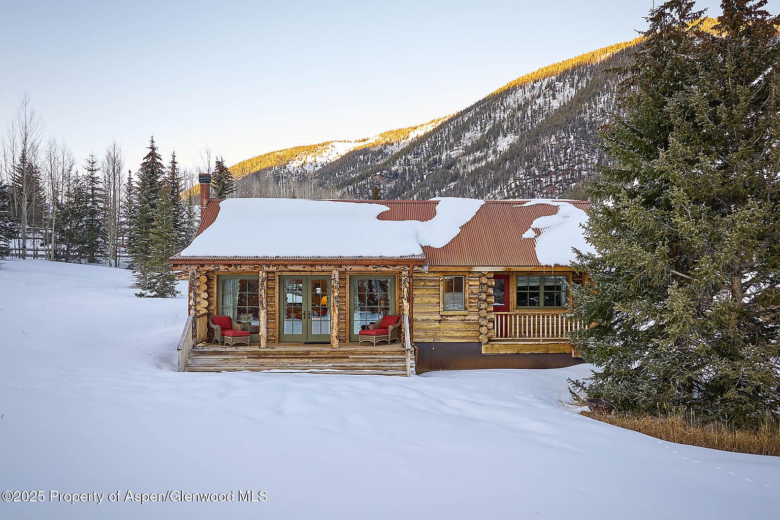 This image showcases the front exterior of a charming log cabin nestled in a snowy landscape. The cabin features a rusty metal roof partially covered in snow, a cozy front porch with red chairs, and large windows that invite natural light. The surrounding trees and mountain backdrop create a serene and picturesque setting, perfect for a secluded retreat.
