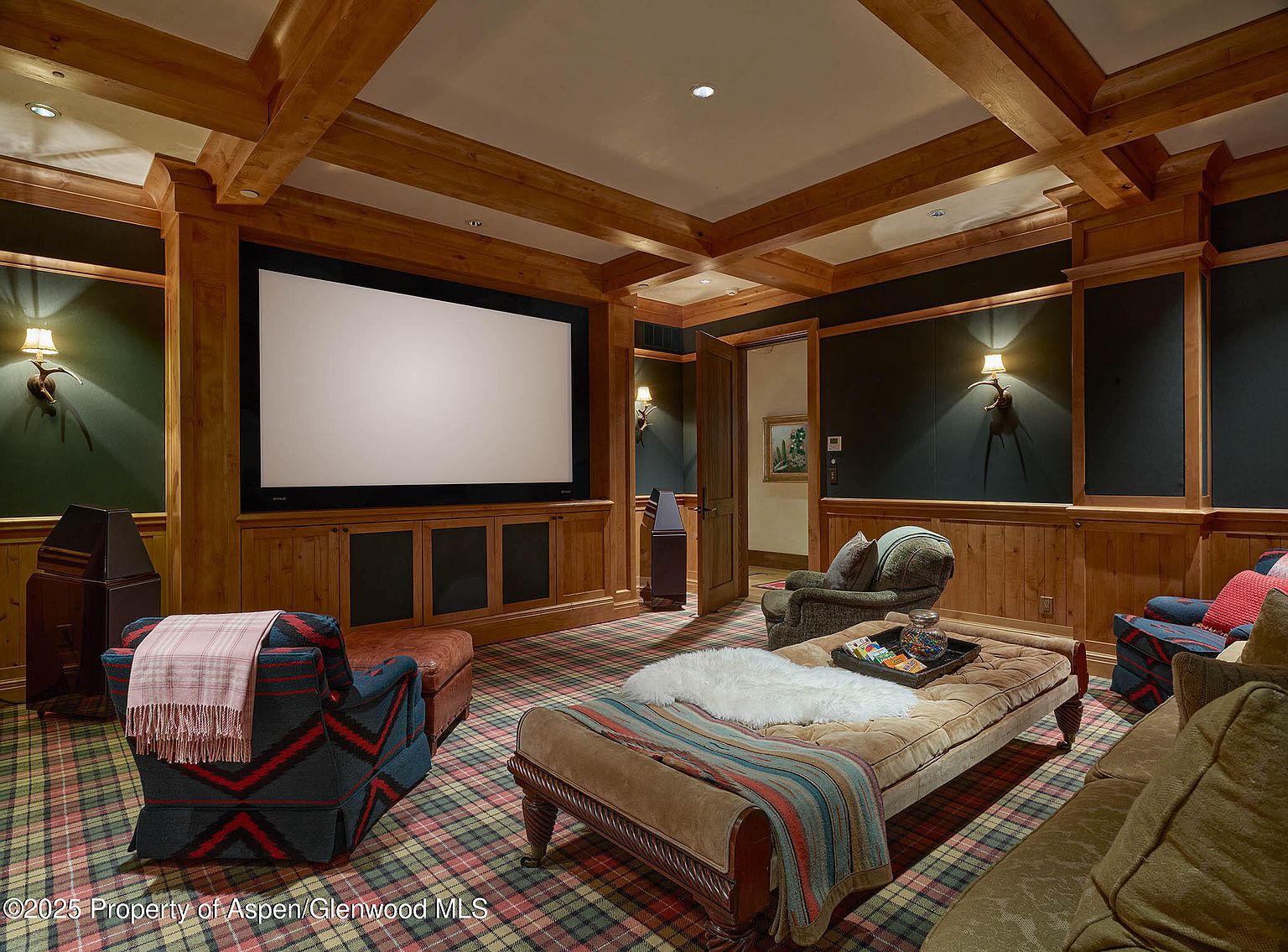 This is an interior shot of a home theater or living room, featuring a large projection screen surrounded by wood paneling and cabinetry. The room is furnished with comfortable seating, including armchairs and a cushioned ottoman, all arranged on a patterned carpet. The space is well-lit by sconces and recessed lighting, creating a cozy and inviting atmosphere.