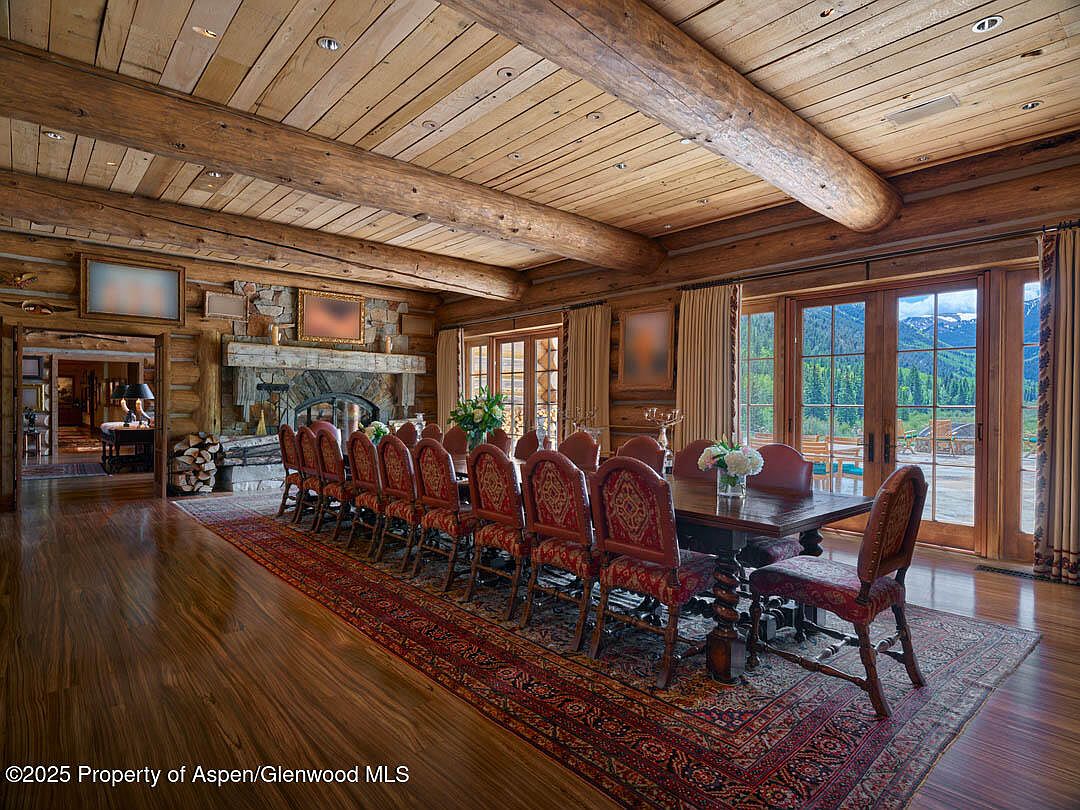 This is an interior shot of a rustic dining room featuring a long wooden table surrounded by ornate chairs with red patterned upholstery. The room has a log cabin aesthetic with exposed wooden beams on the ceiling and walls, a stone fireplace, and large windows offering a view of the mountains. A patterned rug covers the hardwood floor, adding warmth and texture to the space.