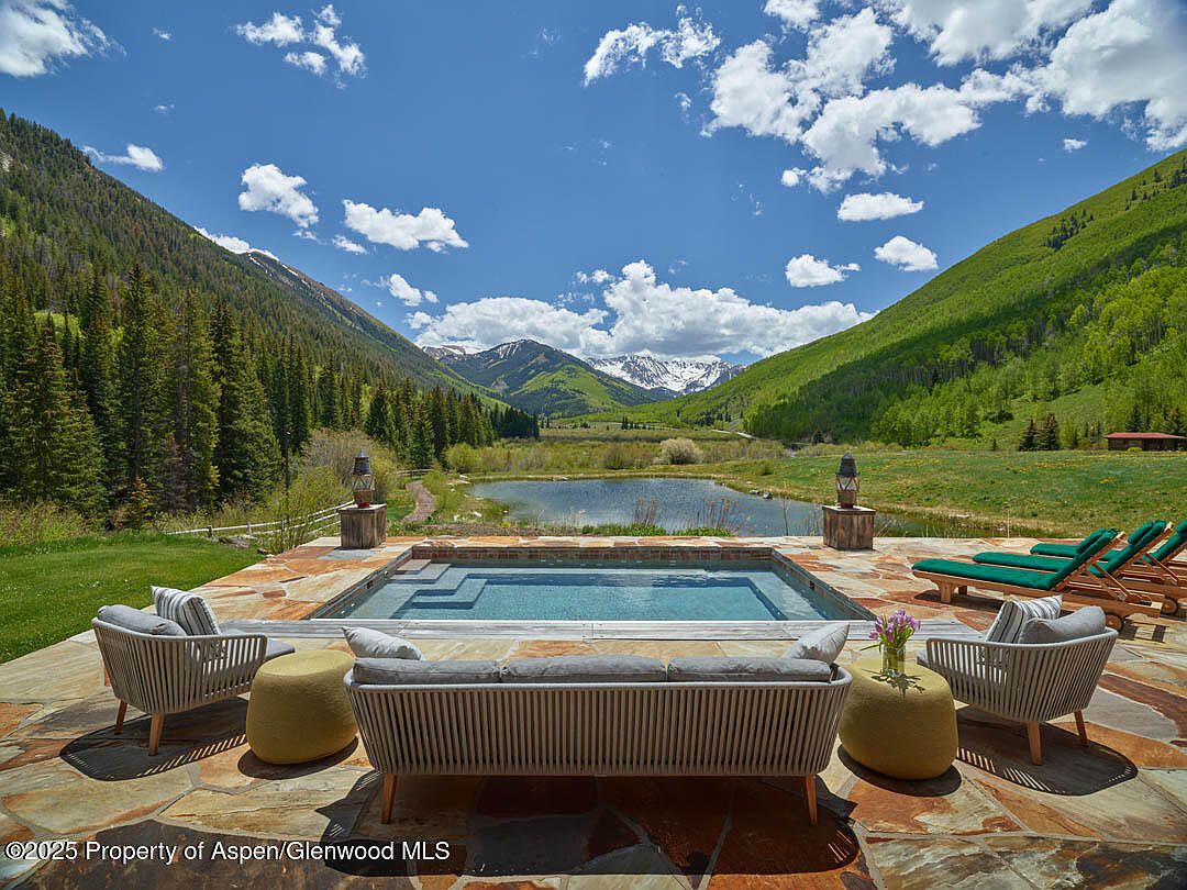 This image showcases a luxurious outdoor pool and spa area with stunning mountain views. The pool is surrounded by a stone patio, comfortable seating, and lounge chairs, creating an inviting space for relaxation. The backdrop features a serene lake and lush green mountains, enhancing the property's appeal.