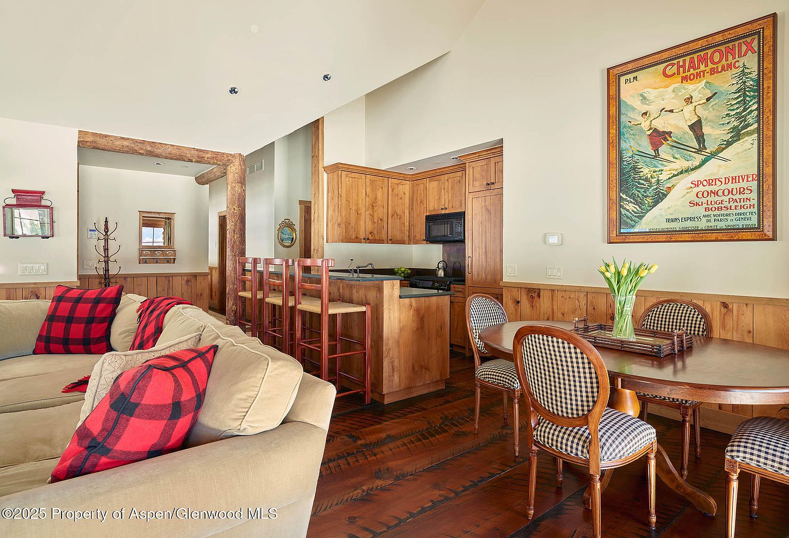 This inviting living space showcases a seamless blend of rustic charm and modern comfort. The room features a cozy sofa adorned with plaid pillows, complemented by a wooden dining table and chairs with patterned upholstery. The adjacent kitchen area boasts wooden cabinetry and a breakfast bar, creating an open and airy atmosphere perfect for entertaining.