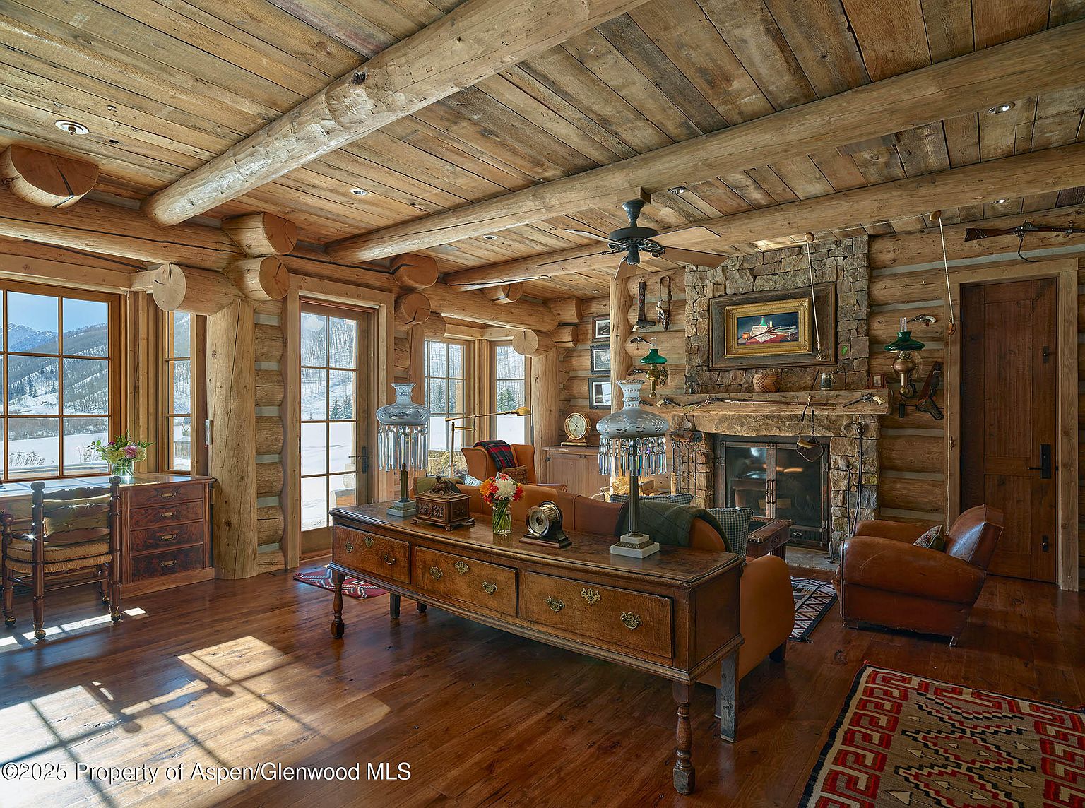 This is a warm and inviting living room with a rustic, log cabin aesthetic. The room features exposed log walls and ceiling beams, a stone fireplace, and hardwood floors. The furniture includes leather armchairs, a wooden console table, and a patterned area rug, creating a cozy and luxurious atmosphere.
