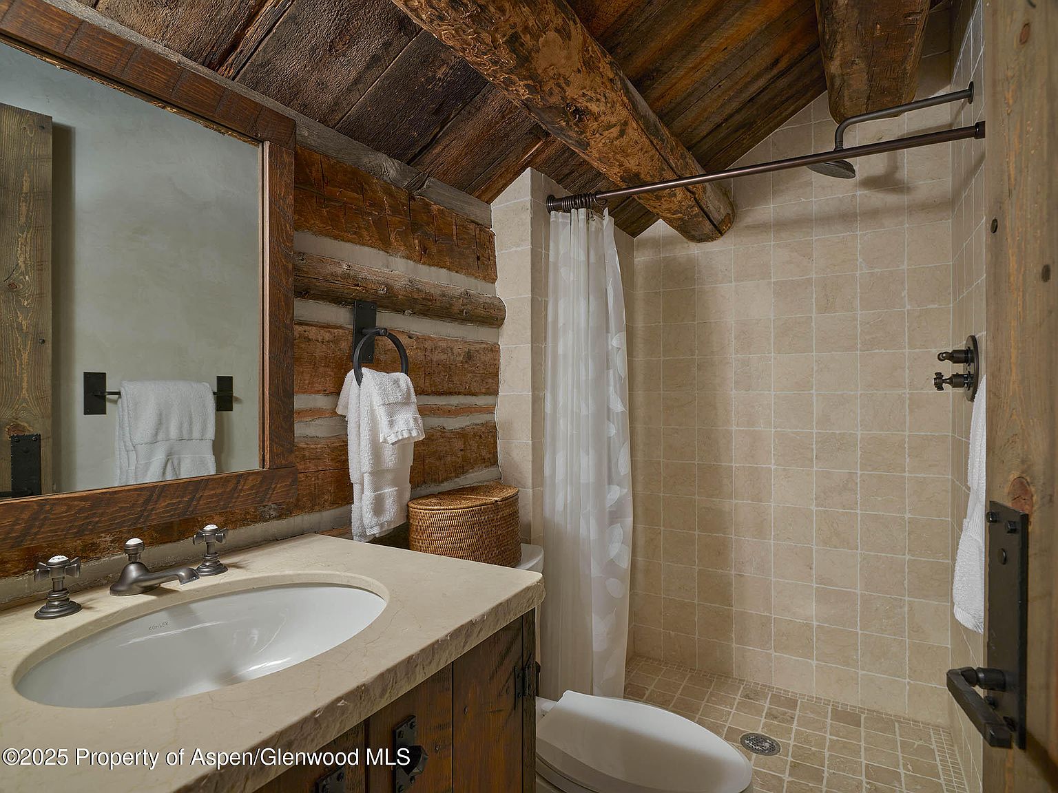This bathroom features a rustic design with log walls and a wooden ceiling. A vanity with a stone countertop and a white sink is visible, along with a framed mirror. The shower area has beige tiles and a shower curtain, creating a cozy and natural ambiance.