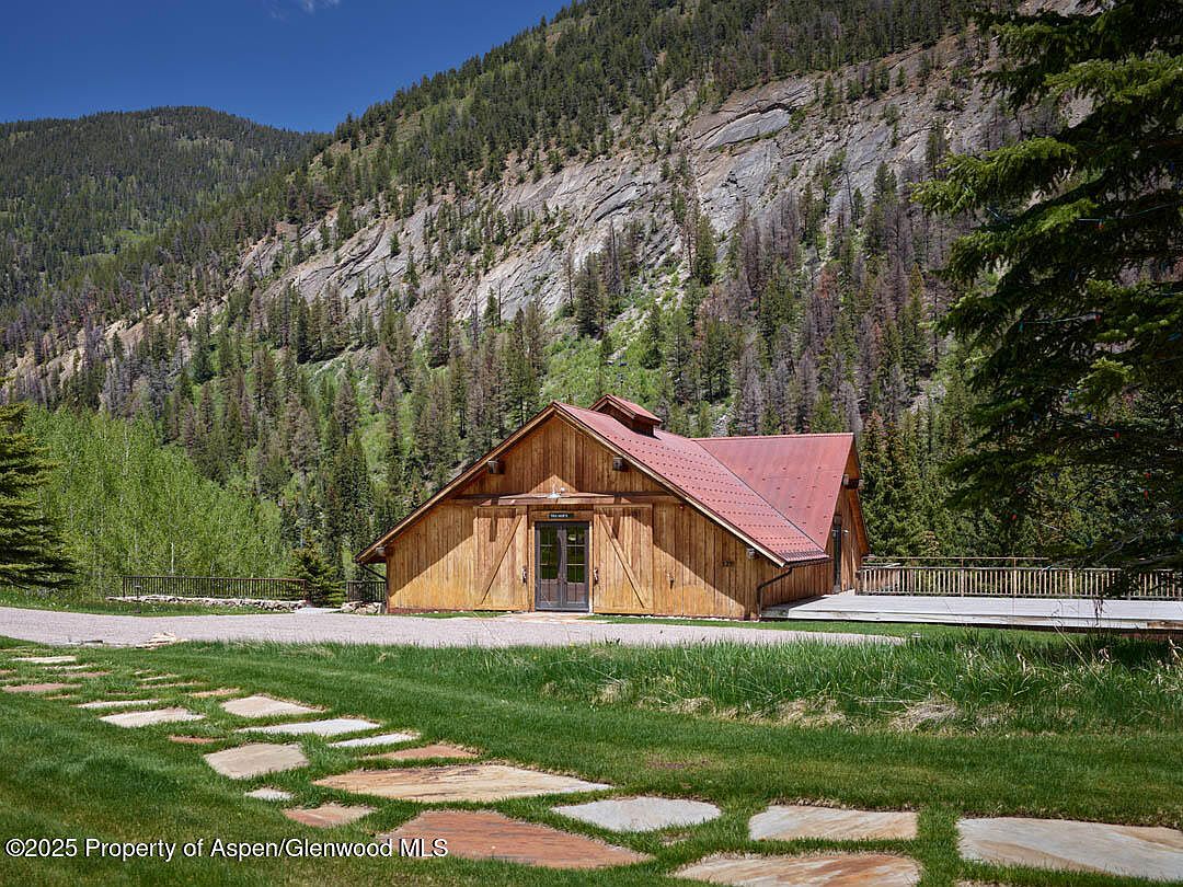The image showcases the front exterior of a rustic barn-style home nestled against a backdrop of a forested mountain. The home features a wooden facade, a red metal roof, and a well-maintained lawn with stone pathways. A wooden deck extends from the side of the house, adding to the property's appeal.