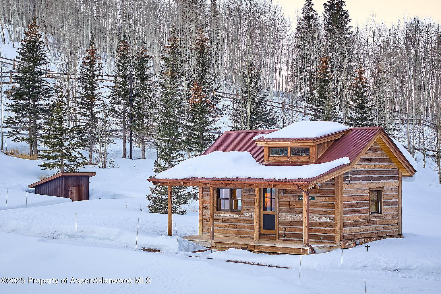 This image showcases the front exterior of a charming log cabin nestled in a snowy landscape. The cabin features a red metal roof, a small porch, and a dormer window, all covered with a layer of fresh snow. Tall evergreen and aspen trees surround the cabin, creating a serene and picturesque winter scene, perfect for a cozy retreat.