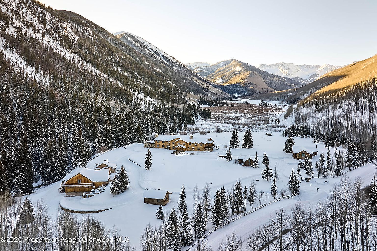 This aerial shot showcases a luxurious mountain estate blanketed in snow. The main lodge, constructed of wood, is surrounded by smaller cabins and dense evergreen forests. The property is nestled in a valley with snow-capped mountains in the background, creating a serene and secluded atmosphere.