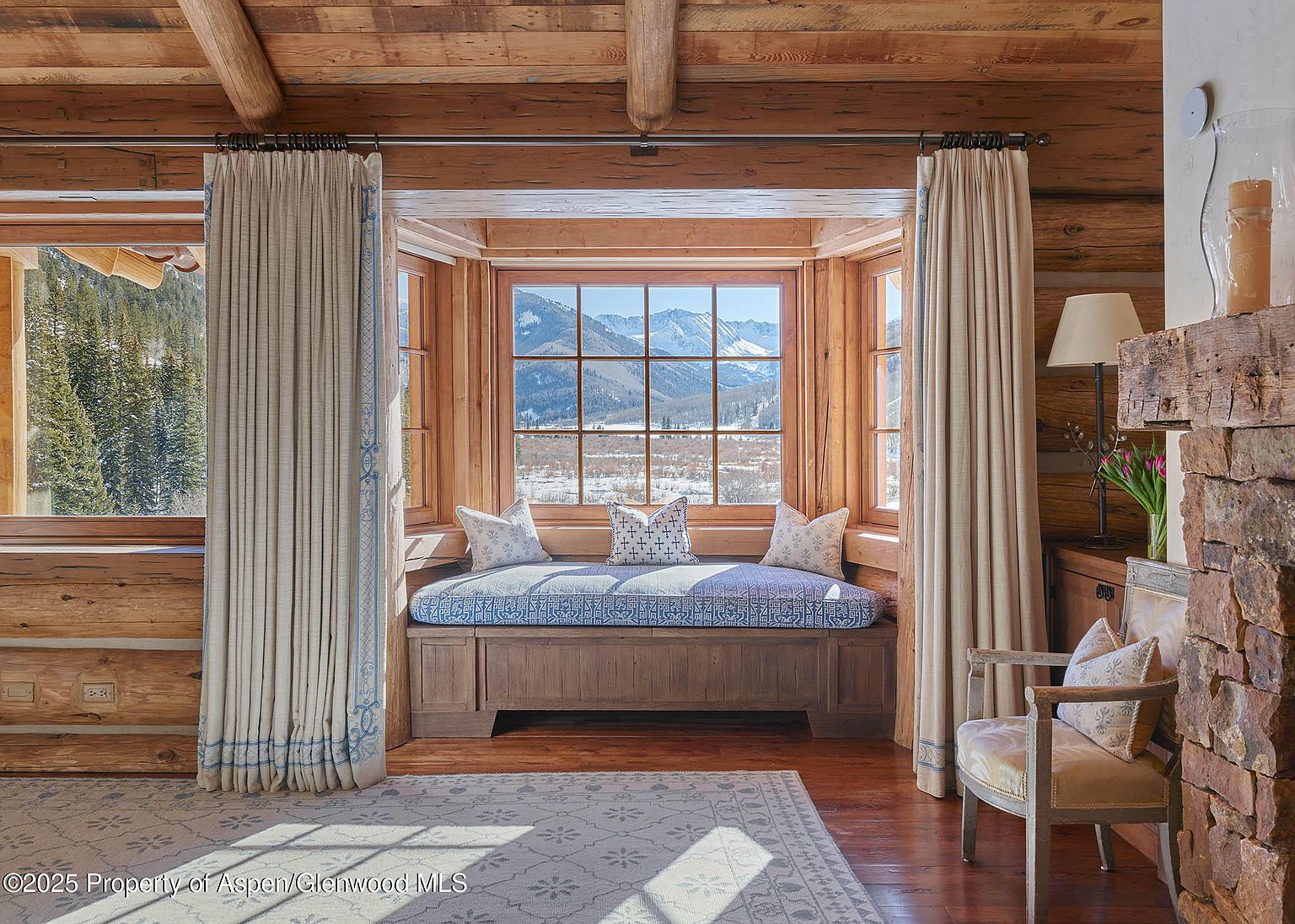 This inviting living room features a cozy window seat with a scenic mountain view, framed by elegant curtains. The room is characterized by its rustic log cabin style, with wooden walls and beams, complemented by a patterned rug and comfortable seating. A stone fireplace adds warmth and character to the space.