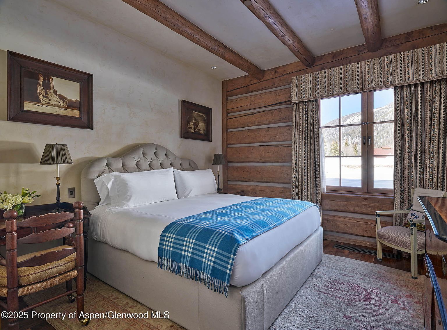 This is a cozy primary bedroom featuring a king-size bed with a tufted headboard and a blue plaid throw. The room has a rustic aesthetic with exposed wooden beams and log walls. Natural light streams in through a window, offering a view of snow-covered mountains.