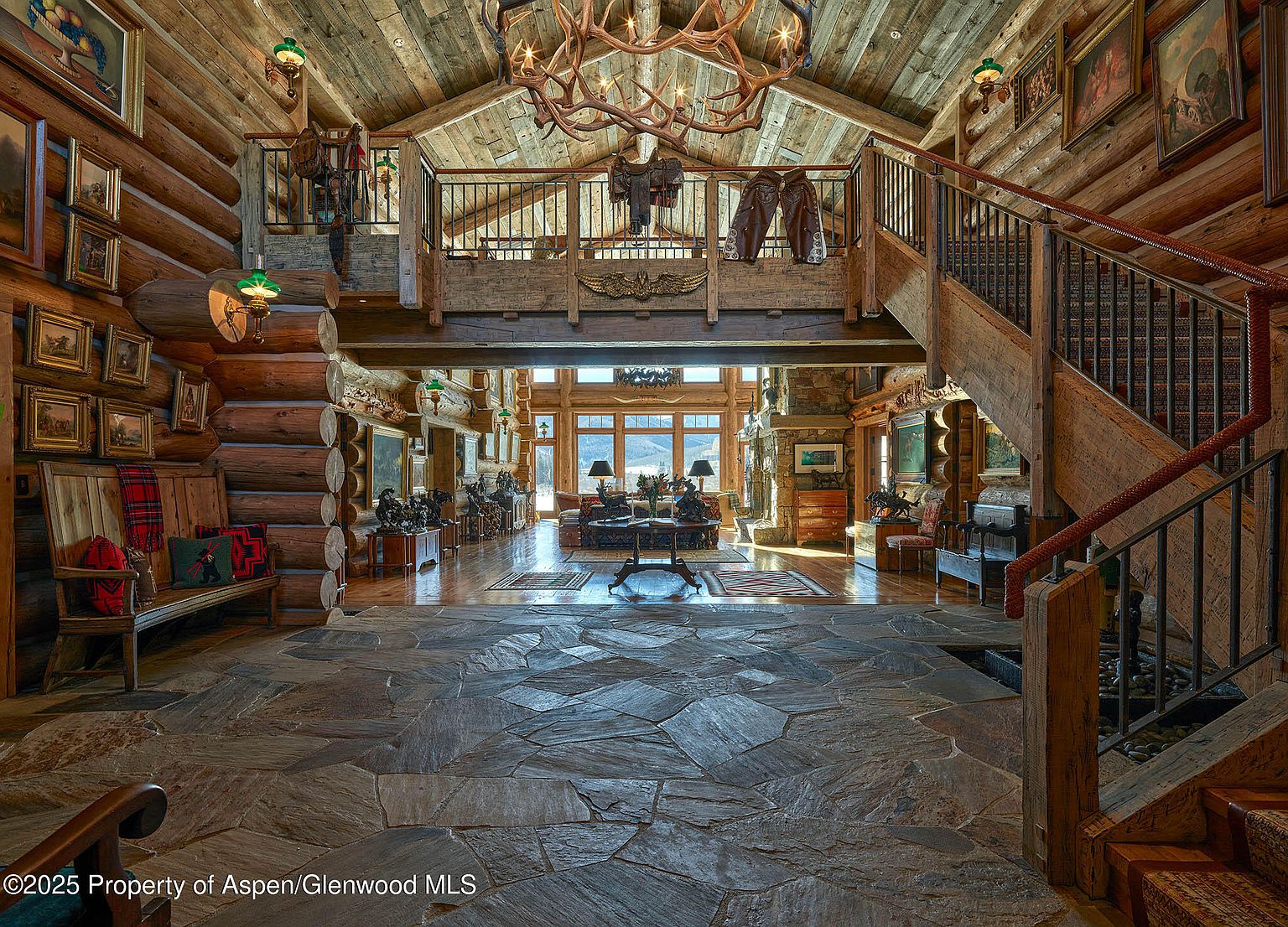 This grand living room features a rustic, lodge-style design with exposed log walls and a high, vaulted ceiling adorned with a striking antler chandelier. A stone floor transitions to hardwood, leading to a bright living area with large windows offering scenic views. The space is furnished with comfortable seating and decorative elements that enhance the room's warm and inviting atmosphere.