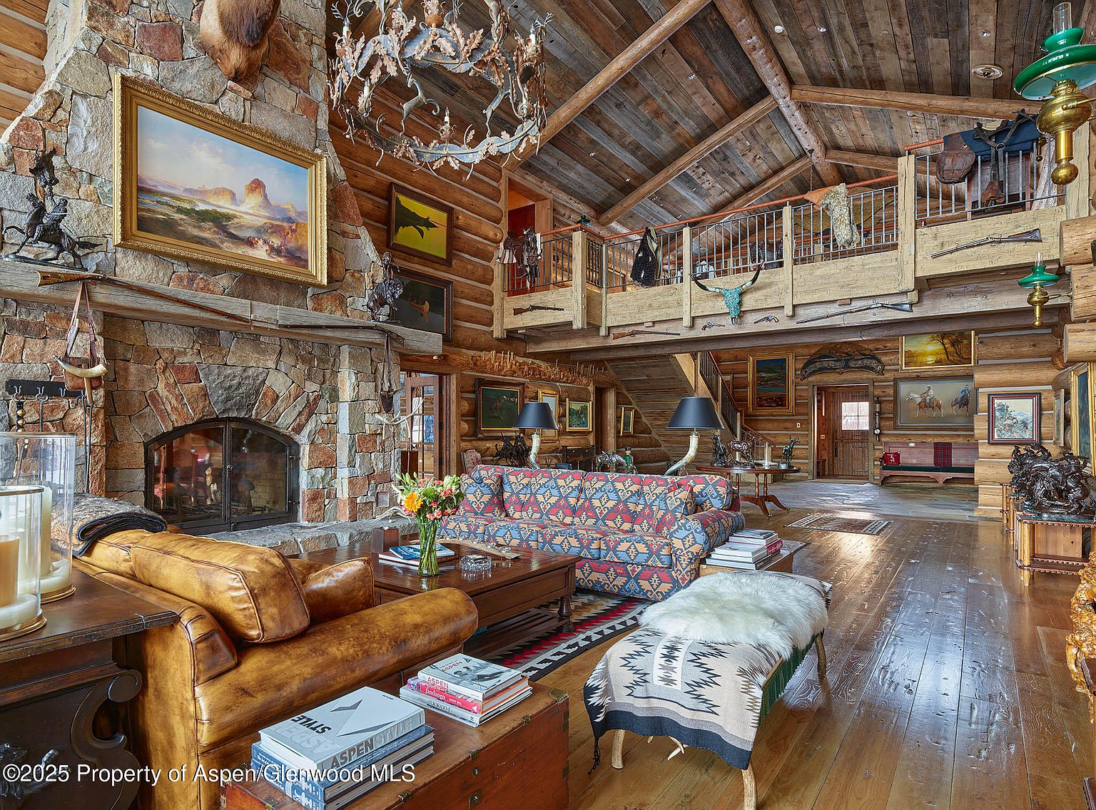 This is a grand living room featuring a rustic, lodge-style design with exposed wooden beams and a stone fireplace. The room is furnished with leather seating, a patterned sofa, and various decorative items such as artwork, sculptures, and mounted animal heads. A mezzanine overlooks the living area, adding to the spacious and luxurious feel of the home.