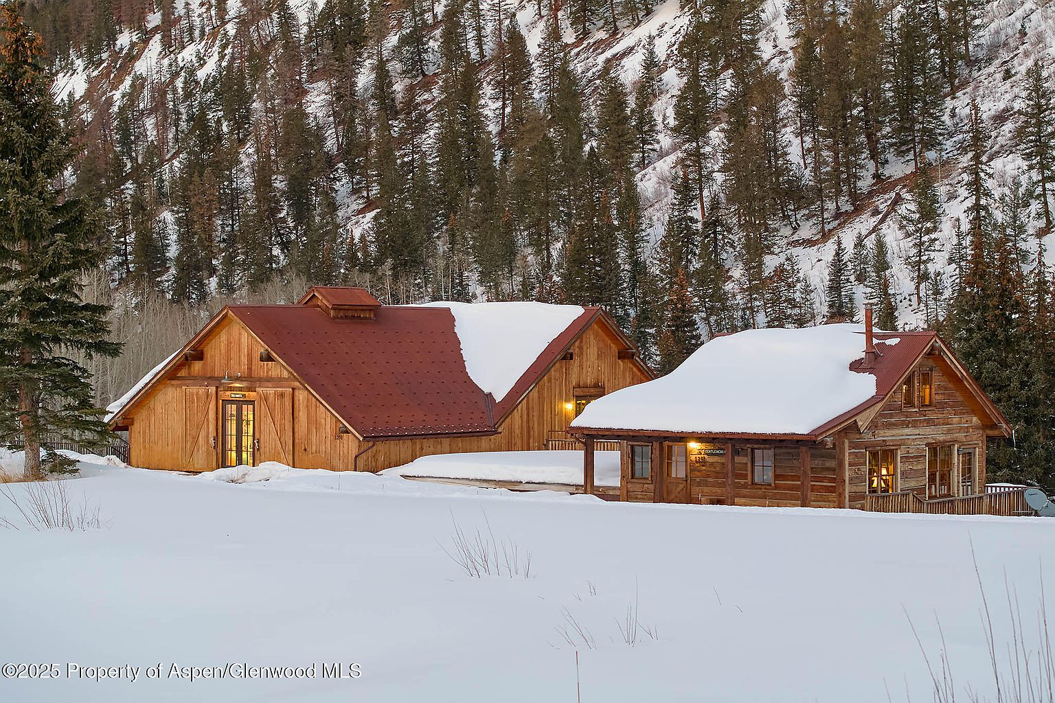 This image showcases the front view of a charming mountain home, likely a cabin or lodge, set against a backdrop of snow-covered trees. The architecture features a combination of wood and a red roof, partially covered in snow, giving it a rustic yet inviting appearance. The scene is serene and picturesque, emphasizing the property's secluded and natural setting.