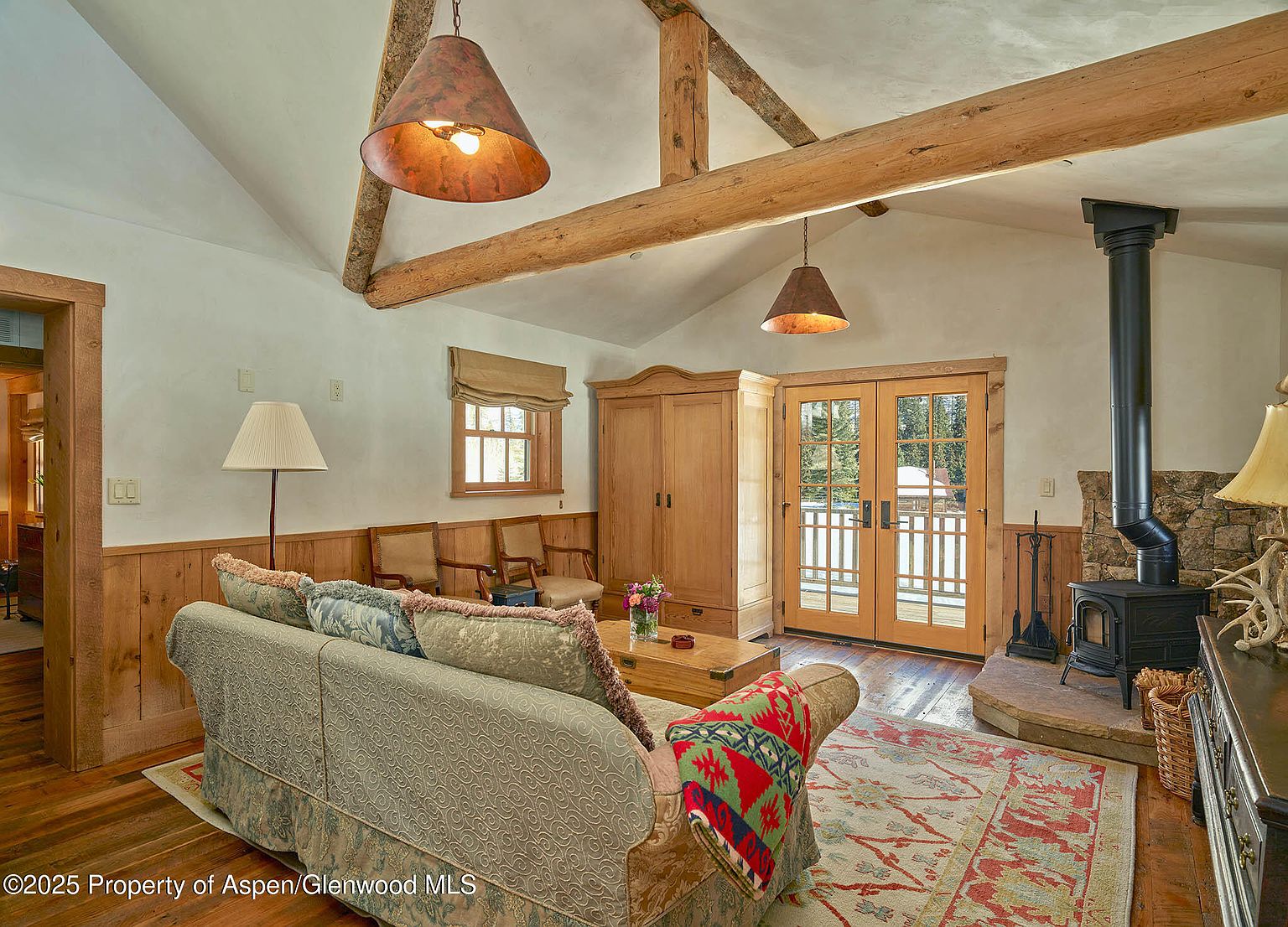 This inviting living room features a vaulted ceiling with exposed wooden beams, creating a rustic and cozy atmosphere. A comfortable sofa is positioned on a patterned rug, complemented by a wood-burning stove in the corner. Natural light streams in through the French doors, enhancing the warm and welcoming ambiance of the space.