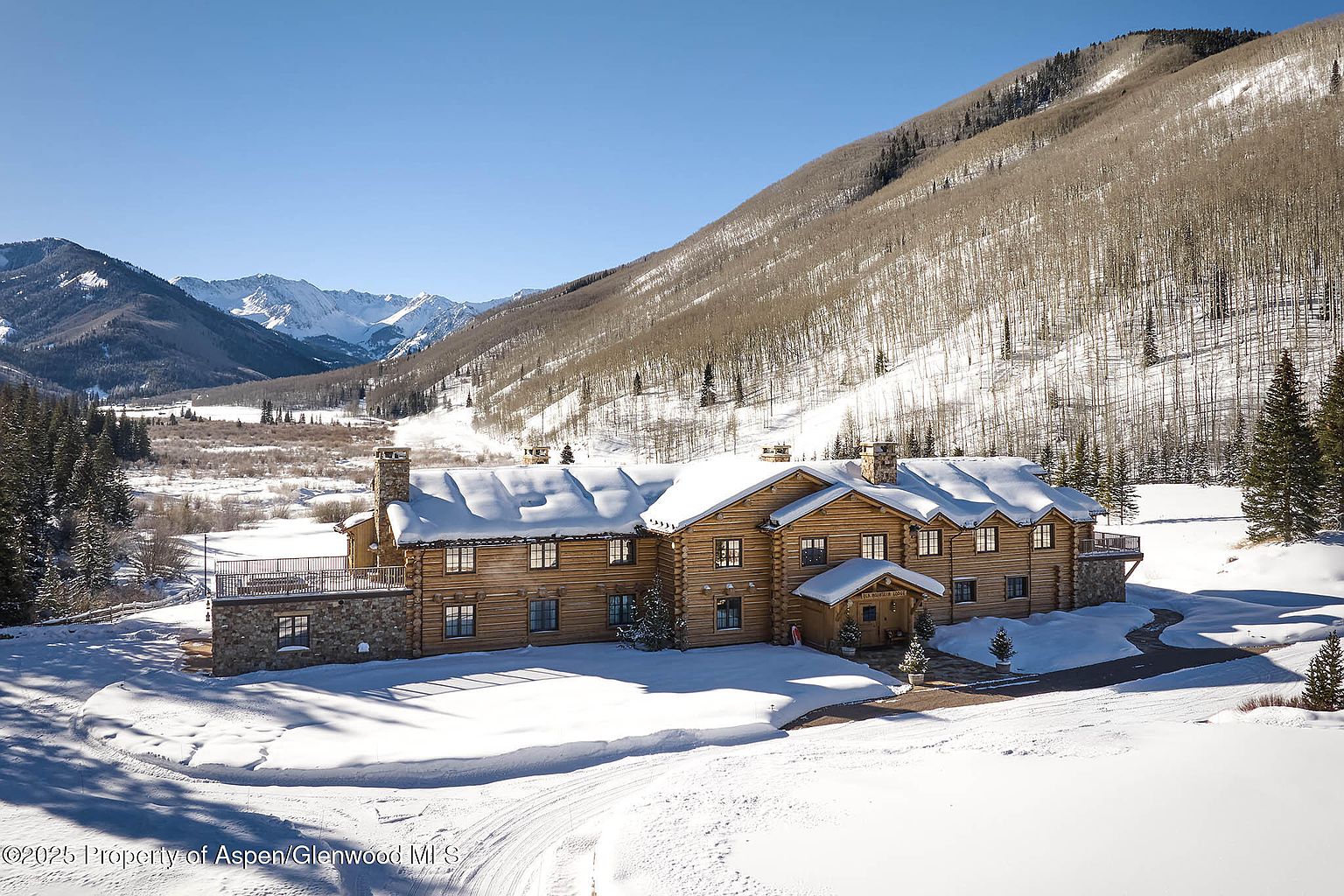 This is a front exterior view of a large, luxurious log cabin home in a snowy, mountainous setting. The home features multiple windows, stone accents, and a snow-covered roof, blending seamlessly with the winter landscape. The driveway is partially cleared, leading to the entrance, and the surrounding area is blanketed in pristine snow, creating a serene and picturesque scene.