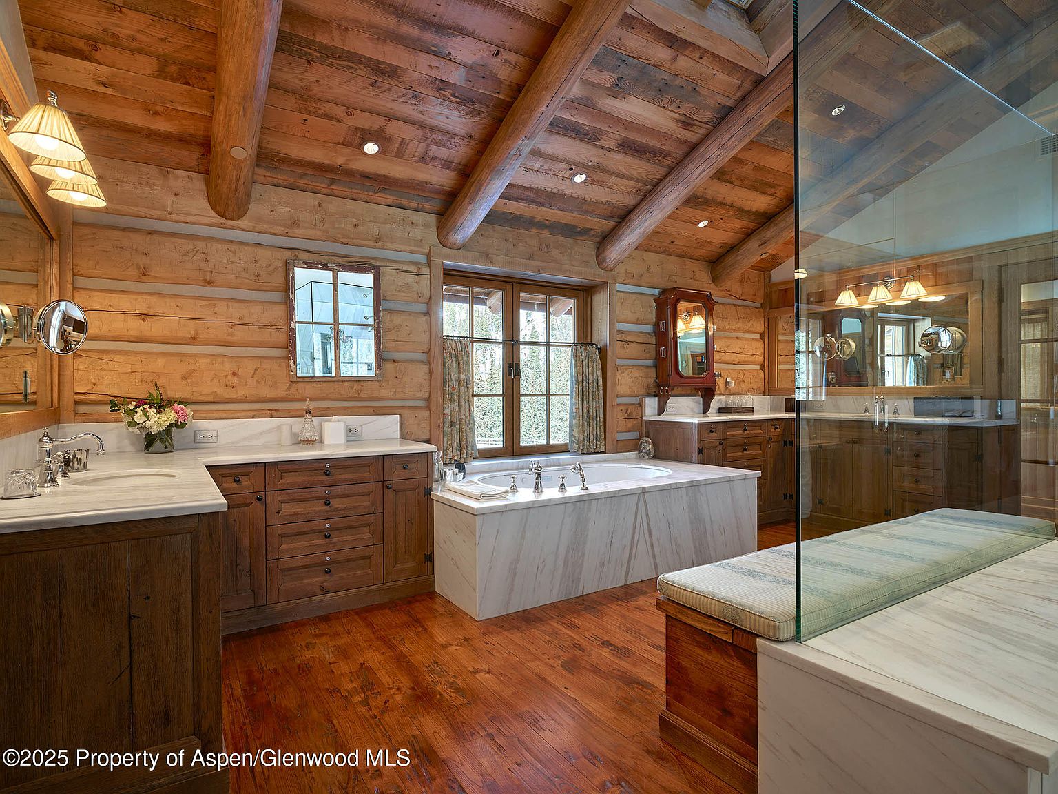 This is a luxurious primary bathroom featuring a rustic log cabin design. The walls and ceiling are constructed from exposed logs, complemented by wooden cabinetry and marble countertops. A large soaking tub sits beneath a window, and a glass-enclosed shower area is visible on the right, creating a spa-like atmosphere.