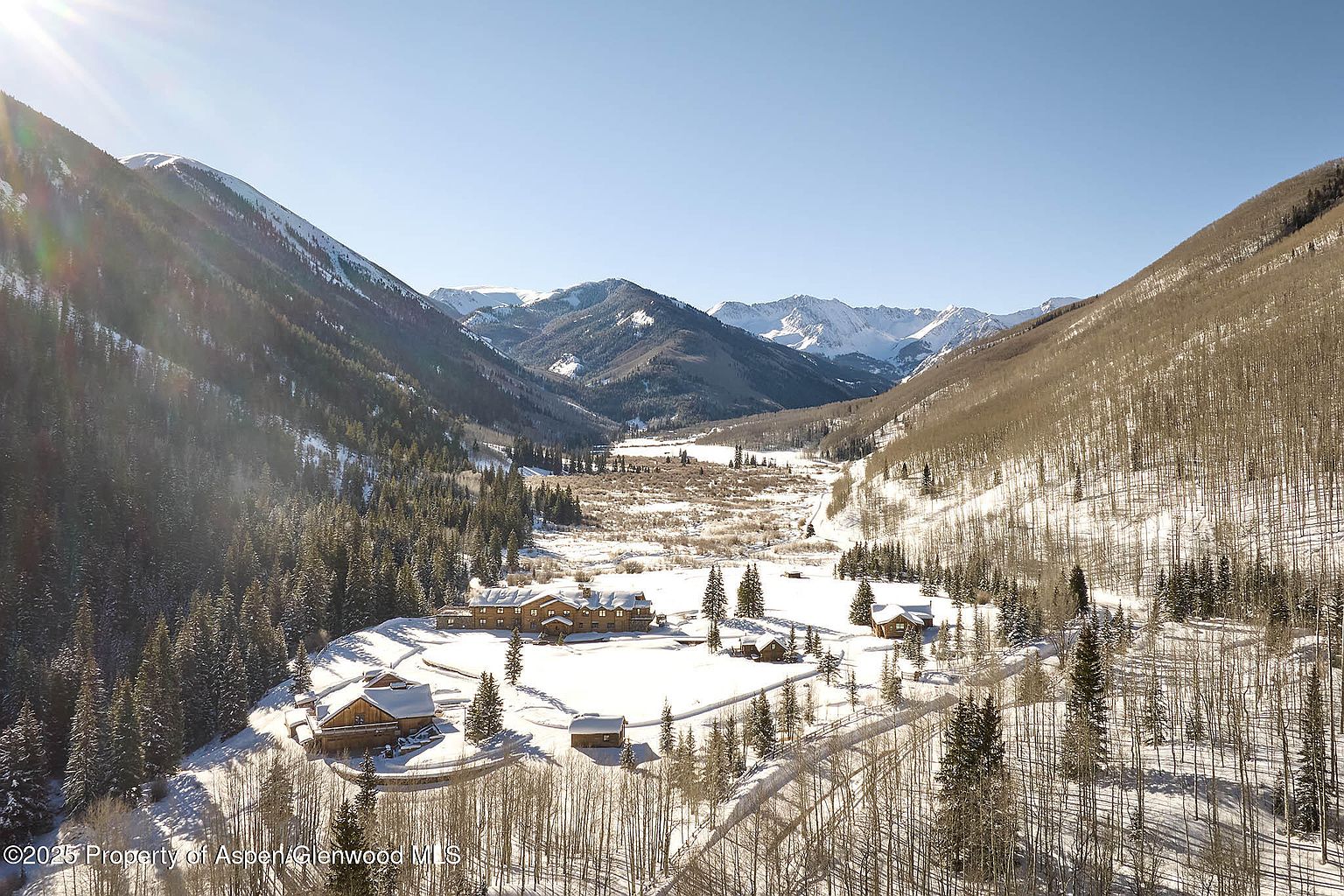 This aerial view showcases a stunning mountain estate nestled in a snow-covered valley. Several log-style buildings are arranged around a central area, surrounded by dense evergreen and deciduous forests. The backdrop features majestic snow-capped mountains under a clear blue sky, creating a serene and secluded atmosphere.