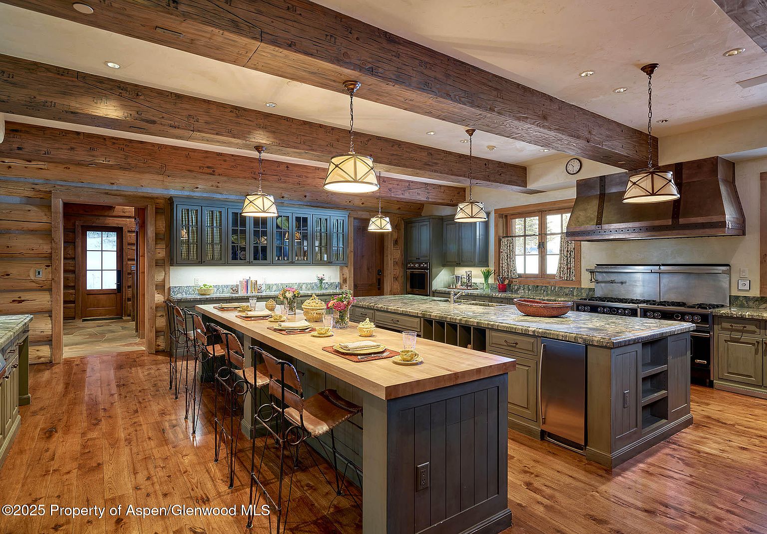 This is a spacious kitchen featuring a large island with seating, wooden countertops, and custom cabinetry. The kitchen is equipped with high-end stainless steel appliances and a professional-grade range. Exposed wooden beams add a rustic touch to the space, complemented by pendant lighting and hardwood floors.