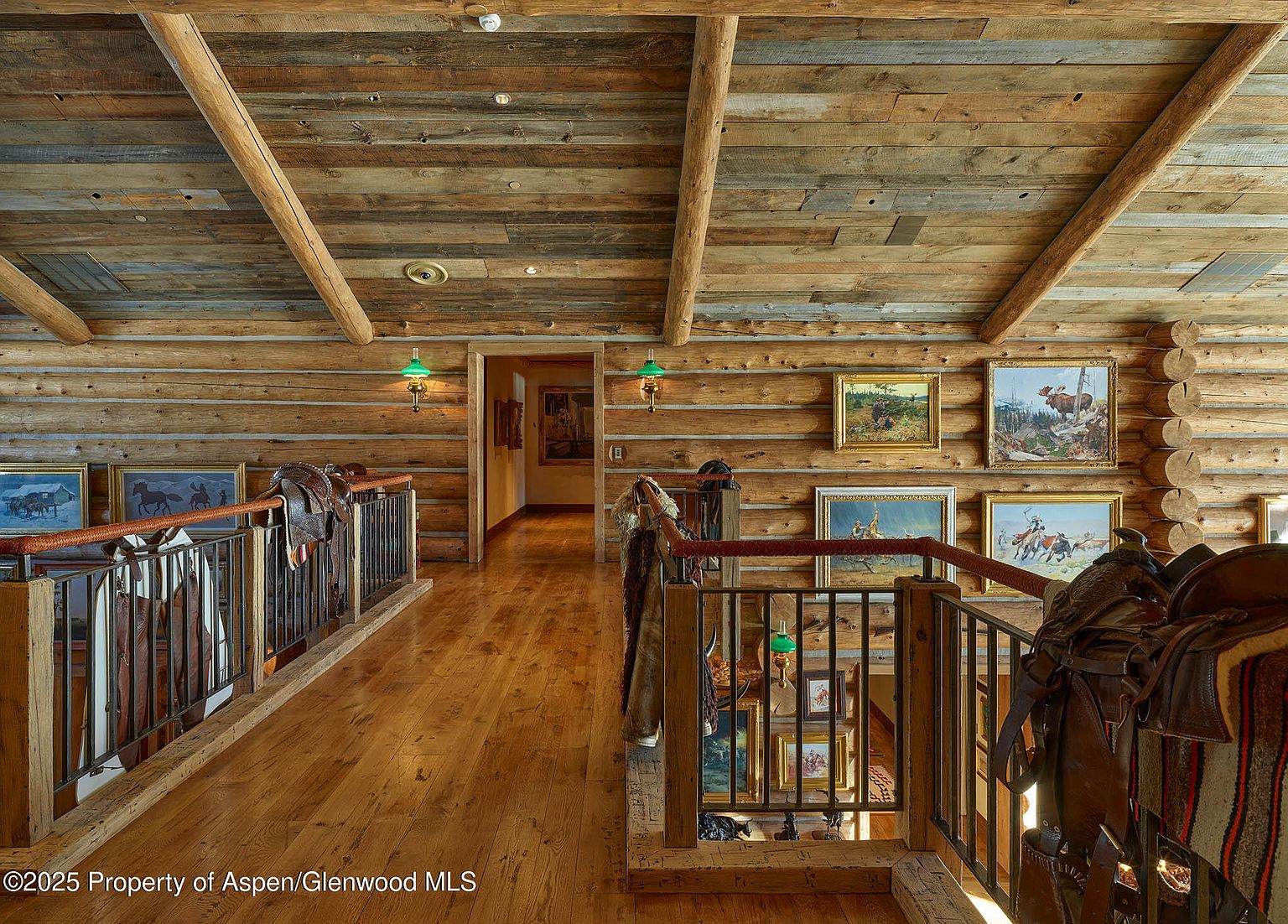 This interior shot showcases a rustic hallway with log walls and a wooden ceiling supported by exposed beams. The hallway features a wooden floor, a railing with metal bars, and artwork displayed on the walls. Saddles are draped over the railing, adding to the western-themed decor.
