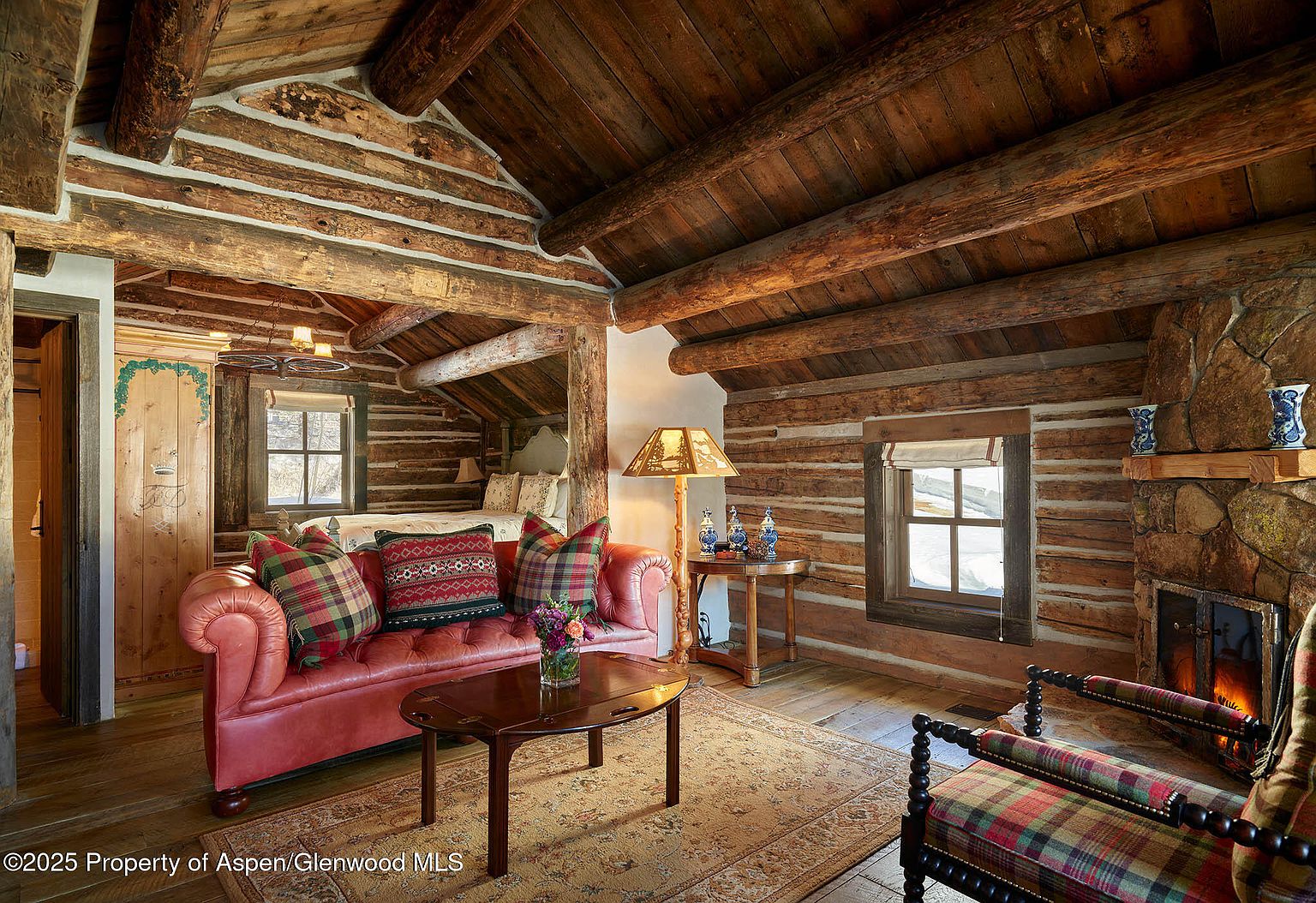 This is a cozy primary bedroom featuring log cabin construction with exposed wooden beams and a stone fireplace. The room is furnished with a red leather sofa, a patterned rug, and a wooden coffee table, creating a warm and inviting atmosphere. Natural light filters through the windows, enhancing the rustic charm of the space.