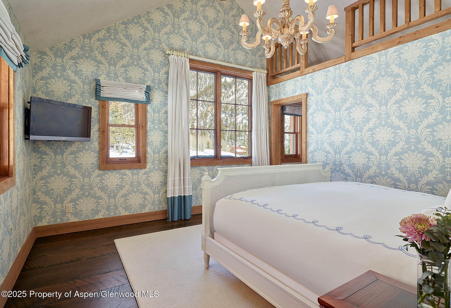 This is a charming bedroom featuring a light blue patterned wallpaper, hardwood floors, and a white bed with blue detailing. Natural light streams in through the wood-framed windows, complemented by a decorative chandelier. The room exudes a cozy and elegant atmosphere, perfect for a guest room.