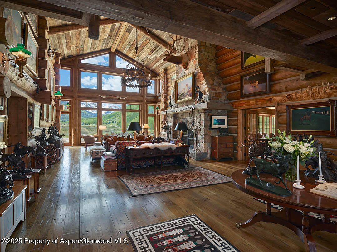 This is a spacious living room with a rustic, lodge-style design. The room features exposed wooden beams, a stone fireplace, and large windows offering a view of the mountains. The decor includes leather furniture, antique tables, and various decorative items, creating a warm and inviting atmosphere.