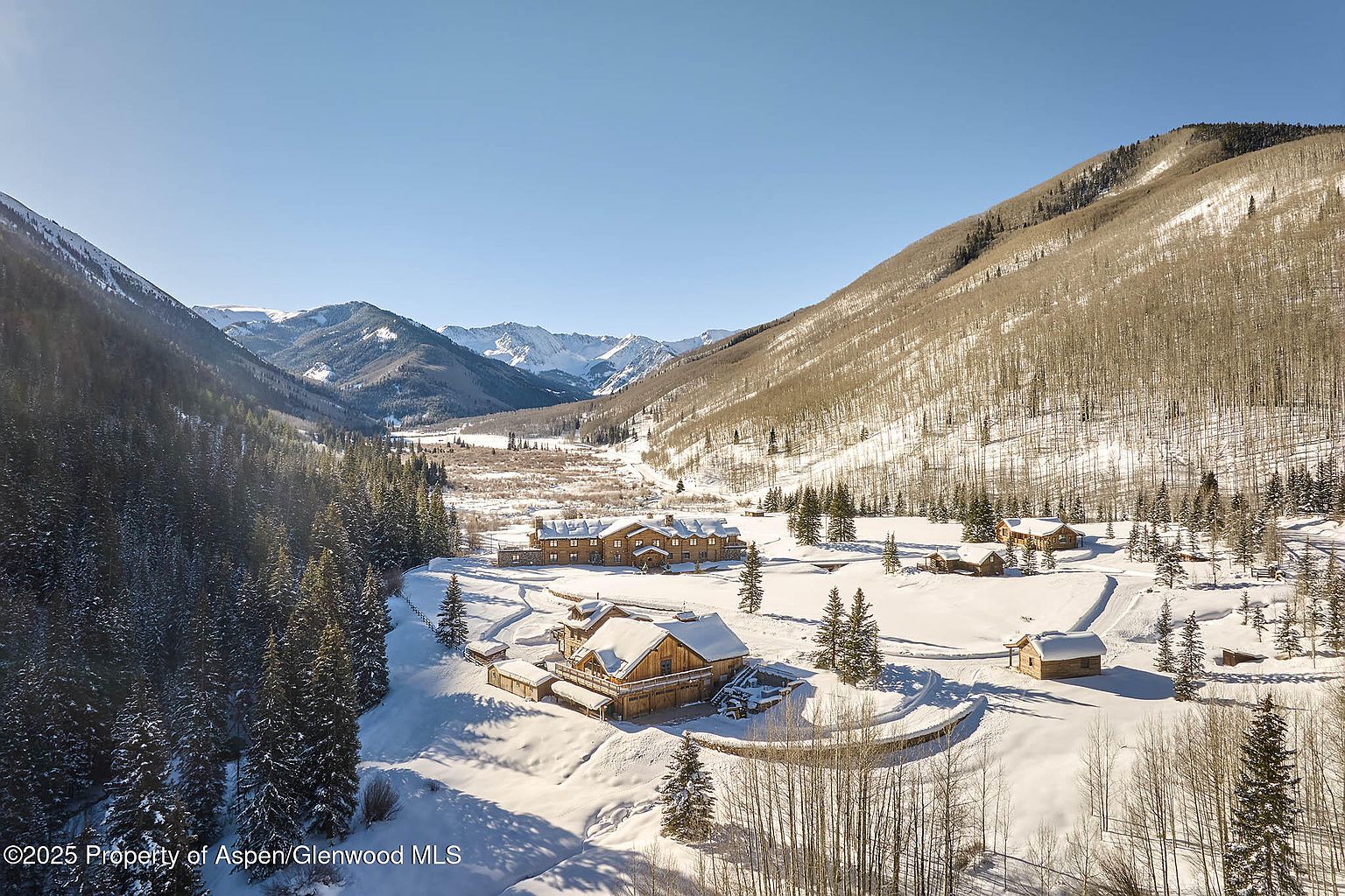 This aerial view showcases a luxurious mountain estate blanketed in snow. The property features multiple log-style buildings, including a main house and several outbuildings, nestled in a valley surrounded by snow-covered mountains and forests. The scene evokes a sense of secluded elegance and winter tranquility.