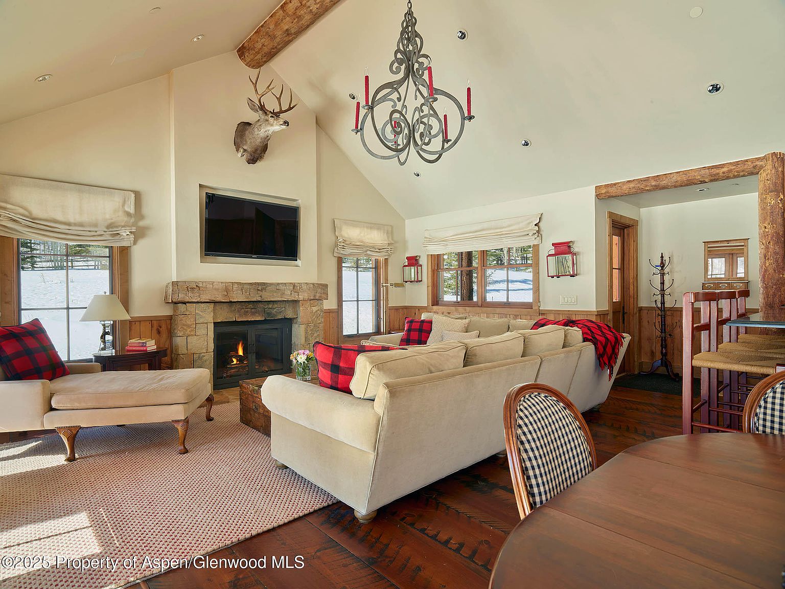 This inviting living room features a stone fireplace with a mounted television above, complemented by a cozy seating arrangement with a large sofa and chaise lounge adorned with plaid accents. The room is illuminated by a unique chandelier and natural light streaming through the windows, creating a warm and rustic atmosphere with exposed wooden beams and hardwood floors. The perspective is from the dining area, showing the living room as a comfortable and stylish space.