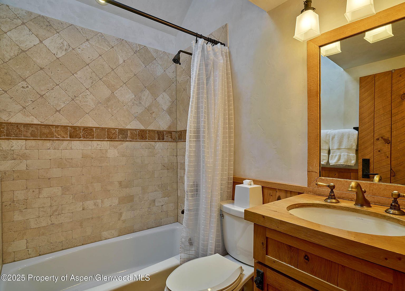 This is a cozy bathroom featuring a bathtub with a shower, a toilet, and a wooden vanity with a sink. The walls are tiled in a neutral tone, and the mirror is framed in wood, complementing the vanity. The overall impression is warm and inviting, with a rustic touch.