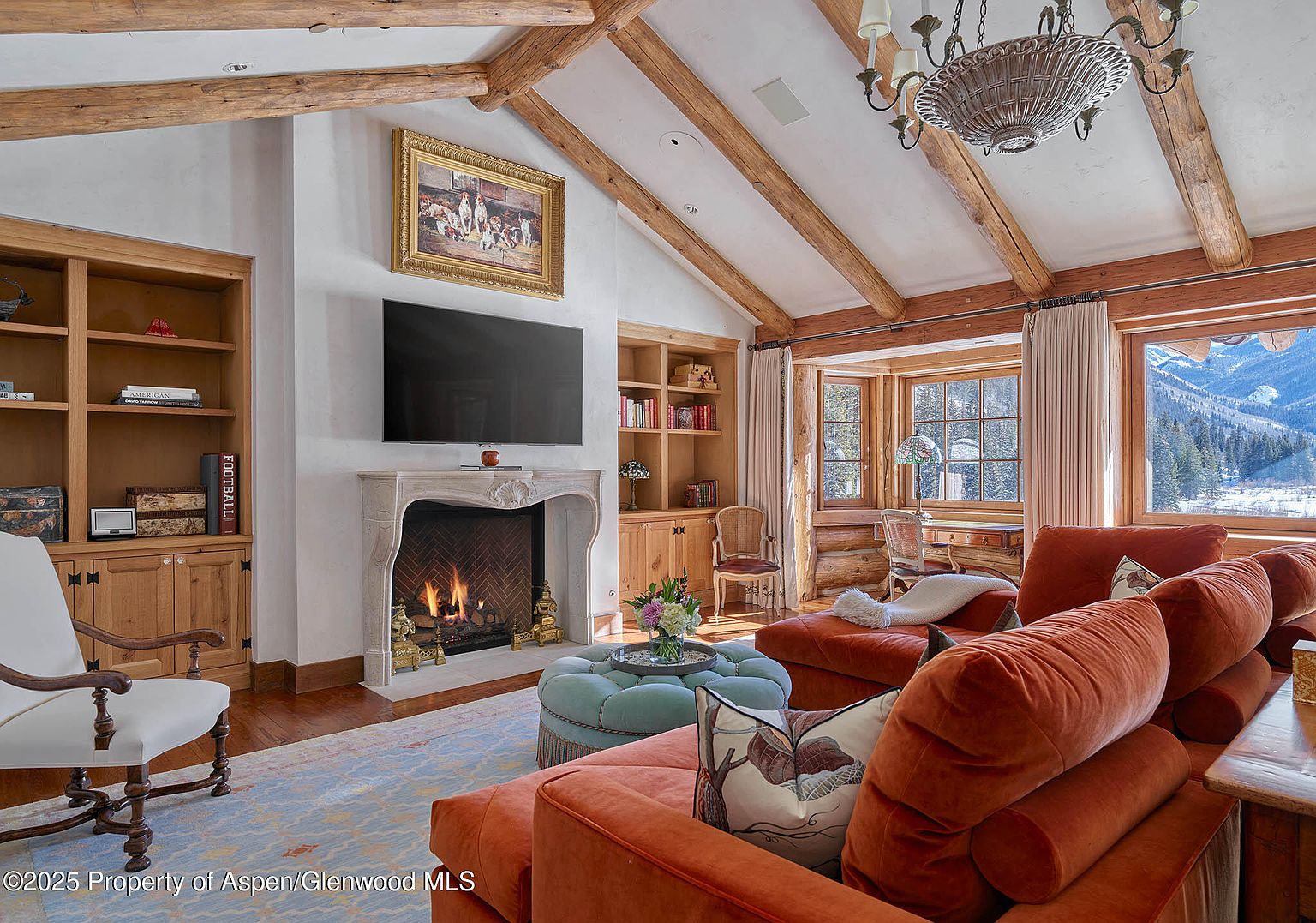 This is an interior shot of a luxurious living room featuring exposed wooden beams, a stone fireplace with a mounted television above, and built-in wooden bookshelves. The room is furnished with comfortable orange sofas and a light blue ottoman, complemented by a patterned rug. Large windows offer a view of a snowy mountain landscape, creating a cozy and inviting atmosphere.