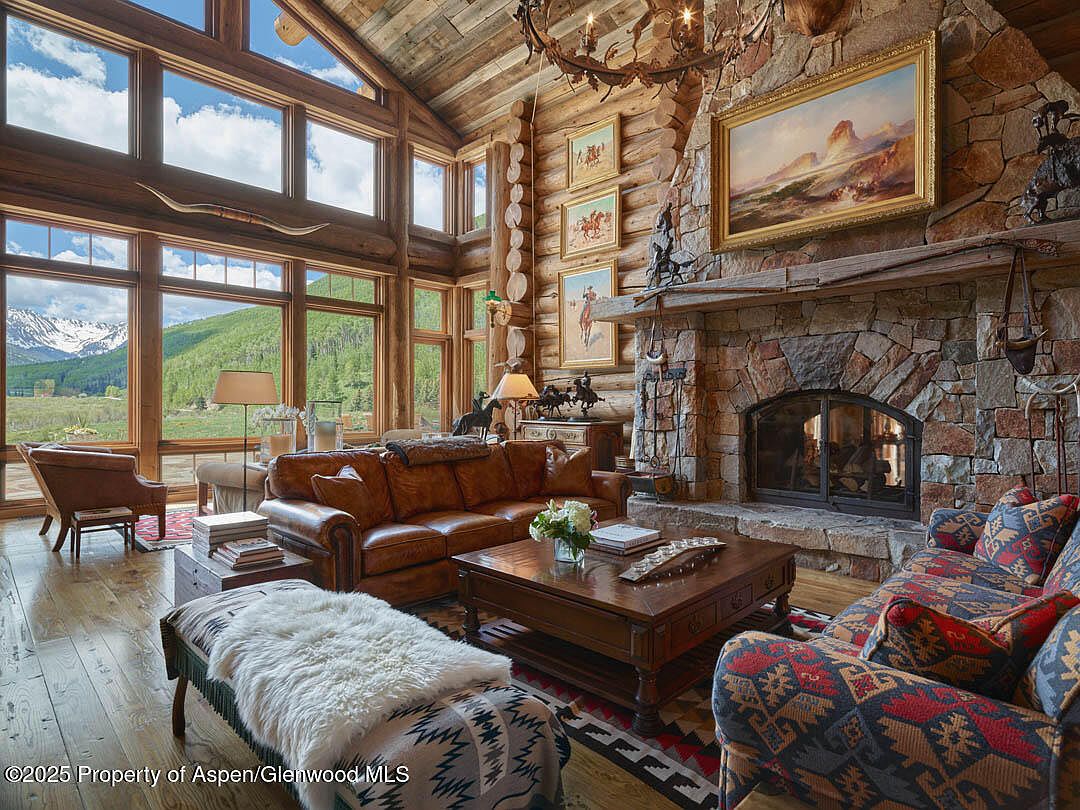 This is an interior shot of a rustic living room featuring a stone fireplace, leather sofa, and large windows with mountain views. The room is decorated with western-themed art and accessories, creating a cozy and luxurious atmosphere. The high ceilings and natural light enhance the spacious feel of the room.