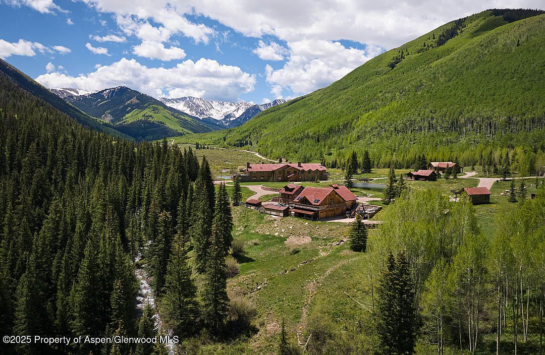 This aerial view showcases a luxurious mountain estate nestled in a lush valley. The property features multiple wooden buildings with red roofs, surrounded by dense evergreen forests and rolling green hills. Snow-capped mountains are visible in the distance, creating a picturesque and serene setting.