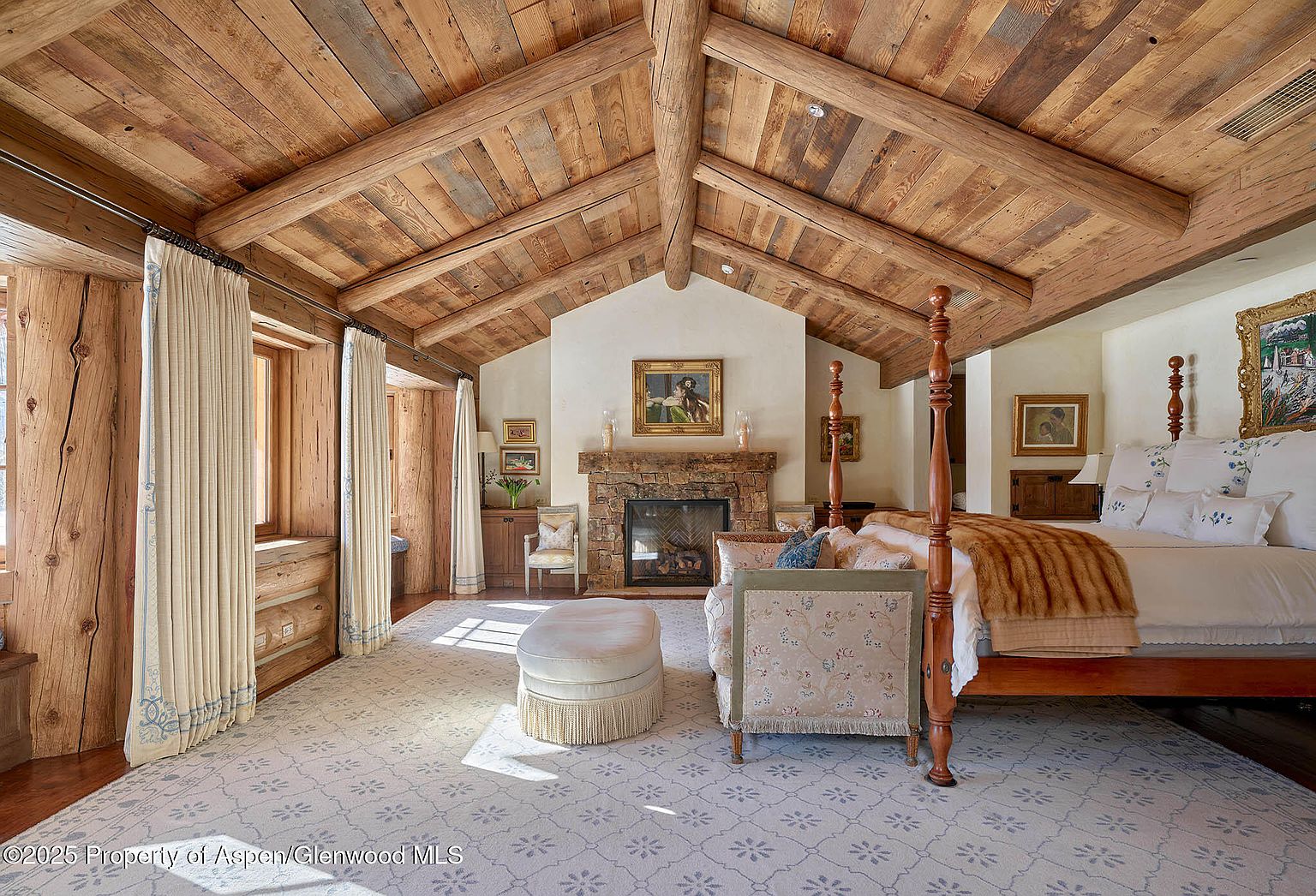 This is a primary bedroom featuring a rustic, luxurious design with a wooden beamed ceiling and a stone fireplace. The room is furnished with a four-poster bed, a patterned rug, and comfortable seating, creating a warm and inviting atmosphere. The natural light enhances the room's cozy and elegant feel, making it a desirable space for relaxation.