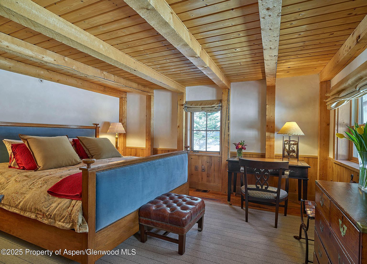 This is a cozy primary bedroom featuring a wooden-beamed ceiling and a large bed with a blue upholstered headboard. The room is decorated with warm tones and includes a desk area with a chair and a dresser. The overall impression is one of rustic elegance and comfort.