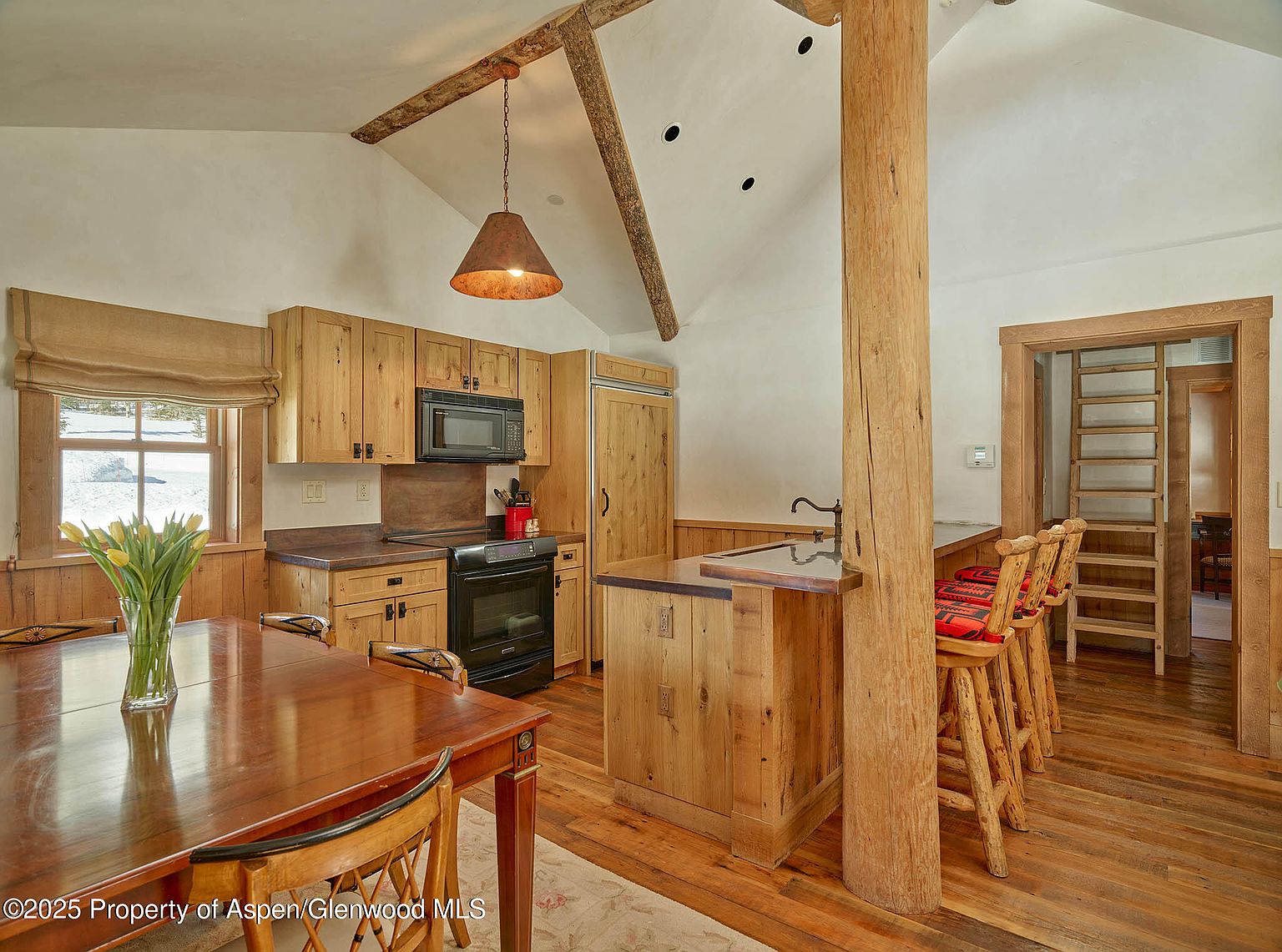 This interior shot showcases a rustic kitchen with wooden cabinetry, countertops, and a central island featuring bar seating. A wooden support beam adds to the rustic charm, while a dining table with a vase of tulips sits in the foreground. The room is well-lit, highlighting the natural wood tones and creating a warm, inviting atmosphere.