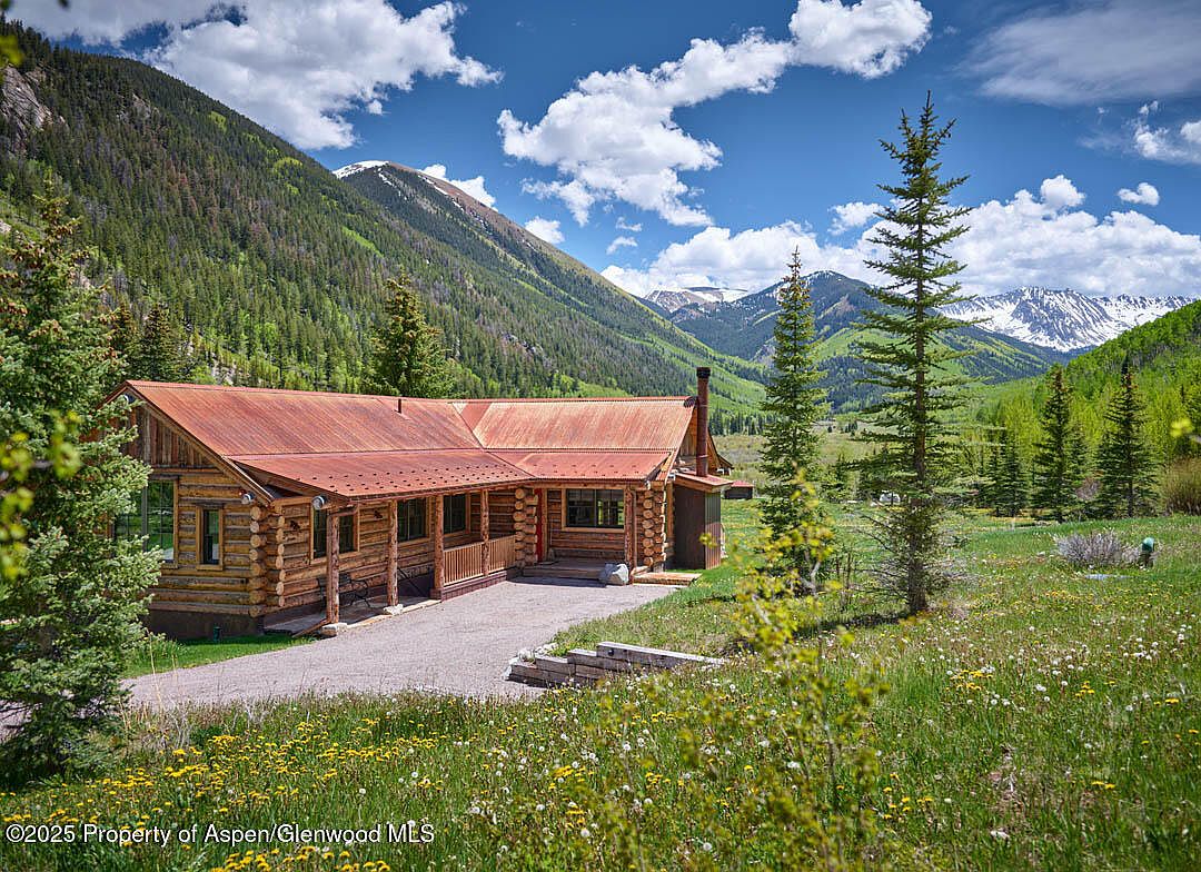 The image showcases a charming log cabin nestled in a picturesque mountain setting. The cabin features a rusty metal roof, a stone foundation, and a welcoming front porch. The surrounding landscape includes lush greenery, wildflowers, and snow-capped mountains in the distance, creating a serene and inviting atmosphere.