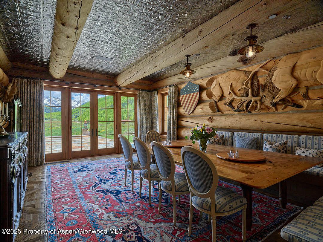 This is an interior shot of a rustic dining room featuring a long wooden table with upholstered chairs and a built-in bench. The room is characterized by log walls, a decorative carved wooden panel, and a patterned ceiling. Large glass doors offer a view of the outdoor landscape, and a patterned rug covers the floor, creating a warm and inviting atmosphere.