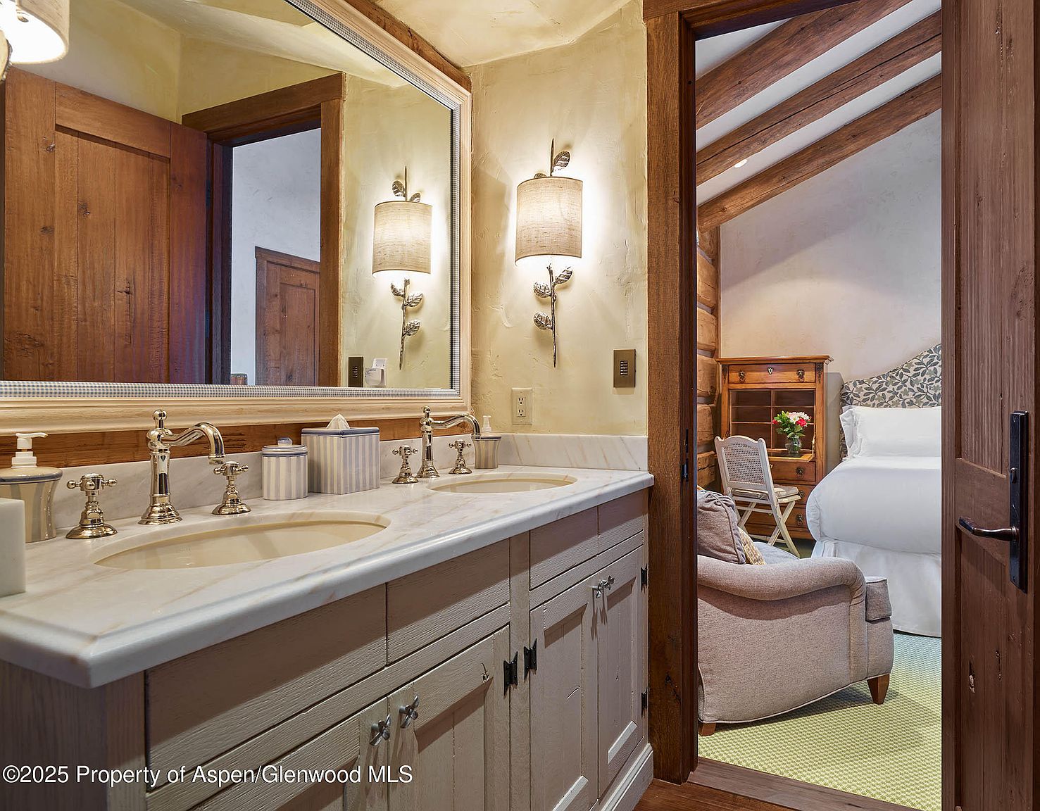 This image showcases a primary bathroom with a double vanity featuring a marble countertop and light-colored cabinetry. The bathroom has rustic-chic elements, including wooden accents and unique sconces. Through the doorway, a glimpse of the bedroom reveals exposed wooden beams and a cozy armchair, suggesting a luxurious and comfortable suite.