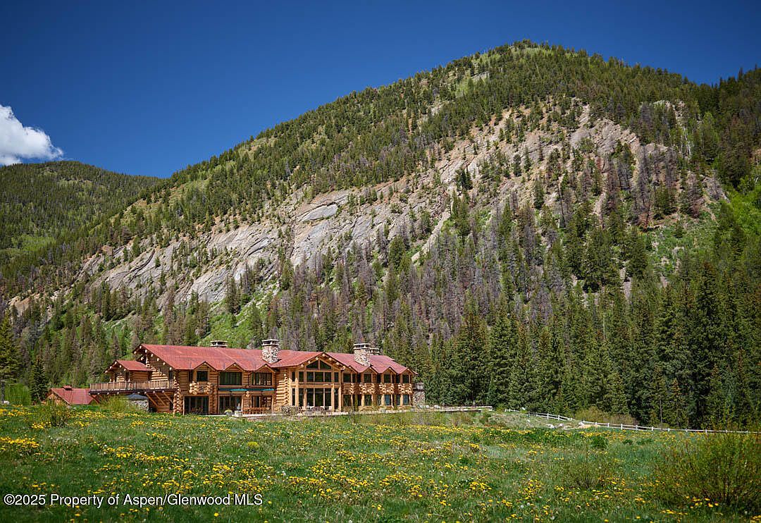 This image showcases the front exterior of a grand log cabin home nestled in a picturesque mountain setting. The home features a red roof, multiple chimneys, and large windows that offer stunning views. The surrounding landscape includes a lush green meadow dotted with yellow wildflowers, creating a serene and inviting atmosphere.