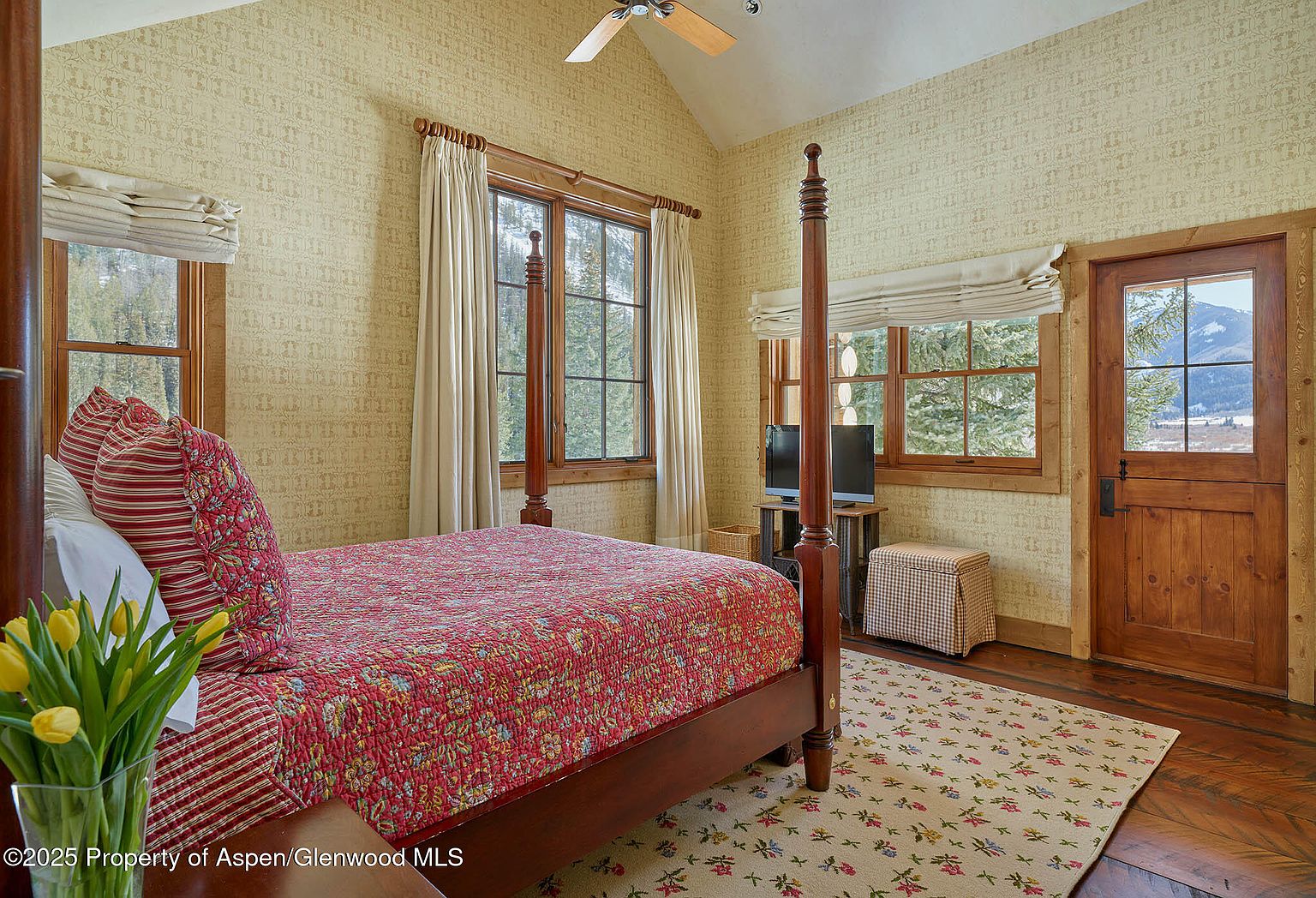 This is a cozy primary bedroom featuring a four-poster bed with a red patterned quilt and matching pillows. The room has patterned wallpaper, wood-framed windows with views of the outdoors, and a floral rug on hardwood floors. The overall style is rustic and inviting, creating a comfortable and relaxing atmosphere.