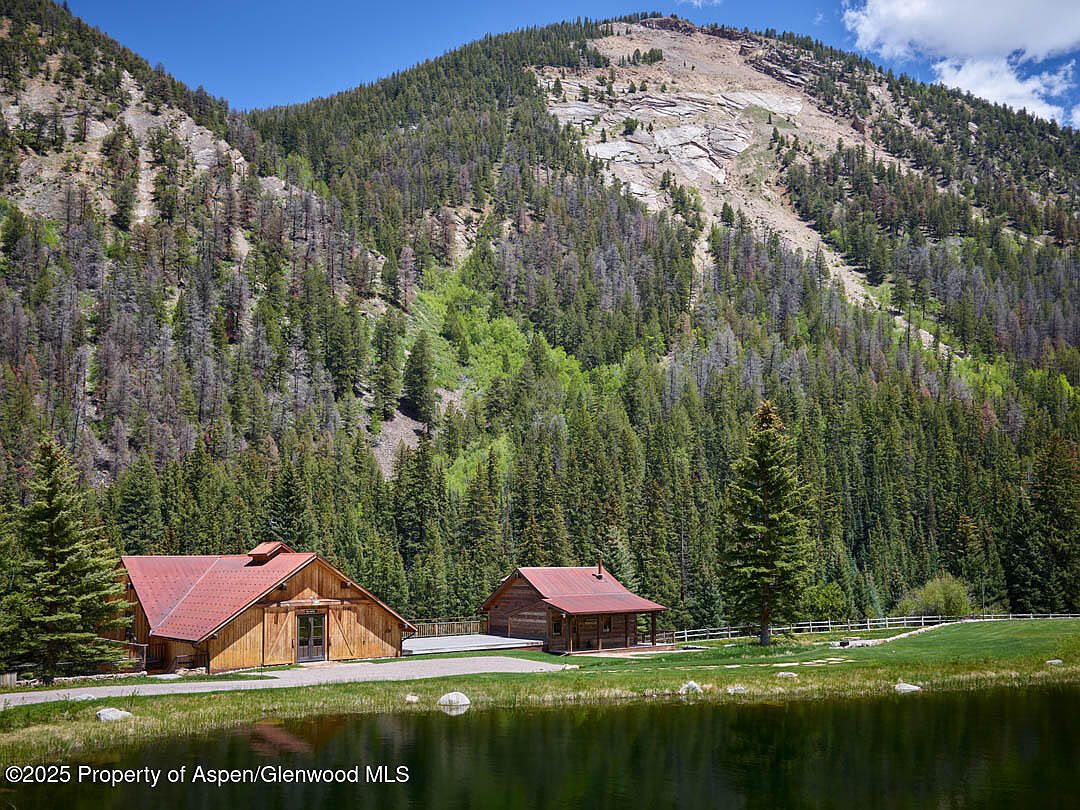 This exterior shot showcases two rustic wooden buildings with red roofs, nestled against a backdrop of lush, forested mountains. The buildings are situated near a calm body of water, reflecting the surrounding landscape. The scene evokes a sense of tranquility and seclusion, highlighting the property's natural setting and potential for a peaceful retreat.