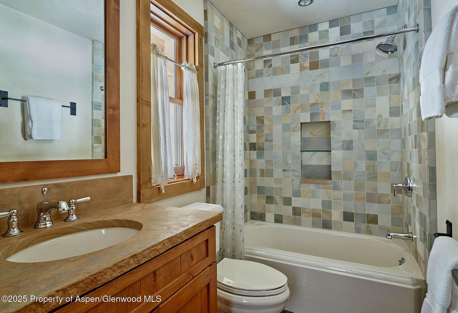 This is a bathroom featuring a wooden vanity with a stone countertop and an oval sink. The shower/tub area is tiled with square tiles in varying shades of gray, beige, and blue, and a shower curtain is partially drawn. A window with sheer curtains provides natural light, and a toilet is visible in the foreground, creating a cozy and functional space.