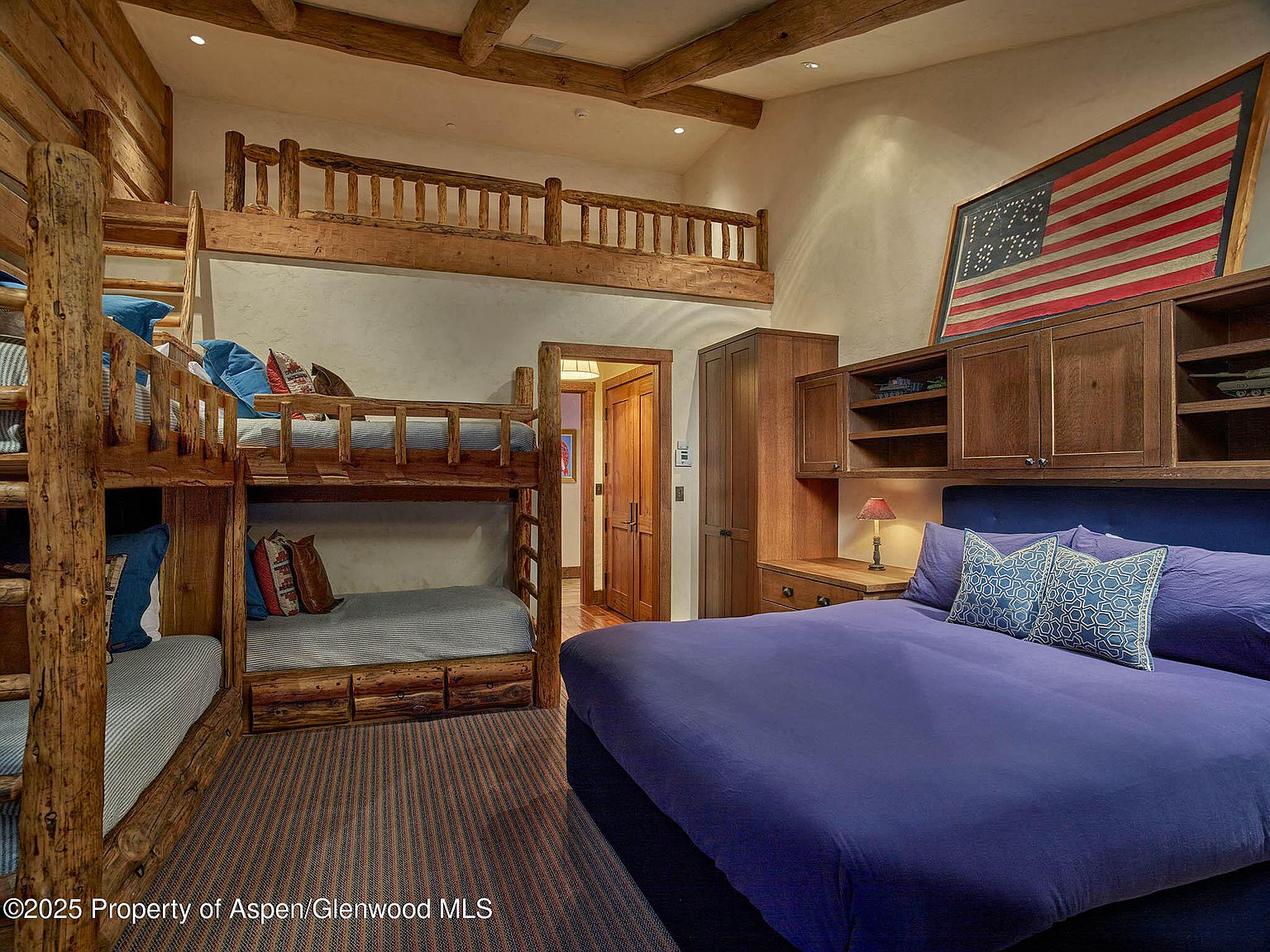 This is an interior shot of a rustic-themed bedroom featuring a unique bunk bed setup with log-style framing and a loft area. The room also includes a large bed with blue bedding, built-in wooden cabinets, and an American flag decoration above the bed. The overall impression is cozy and inviting, with a focus on natural materials and a lodge-like atmosphere.