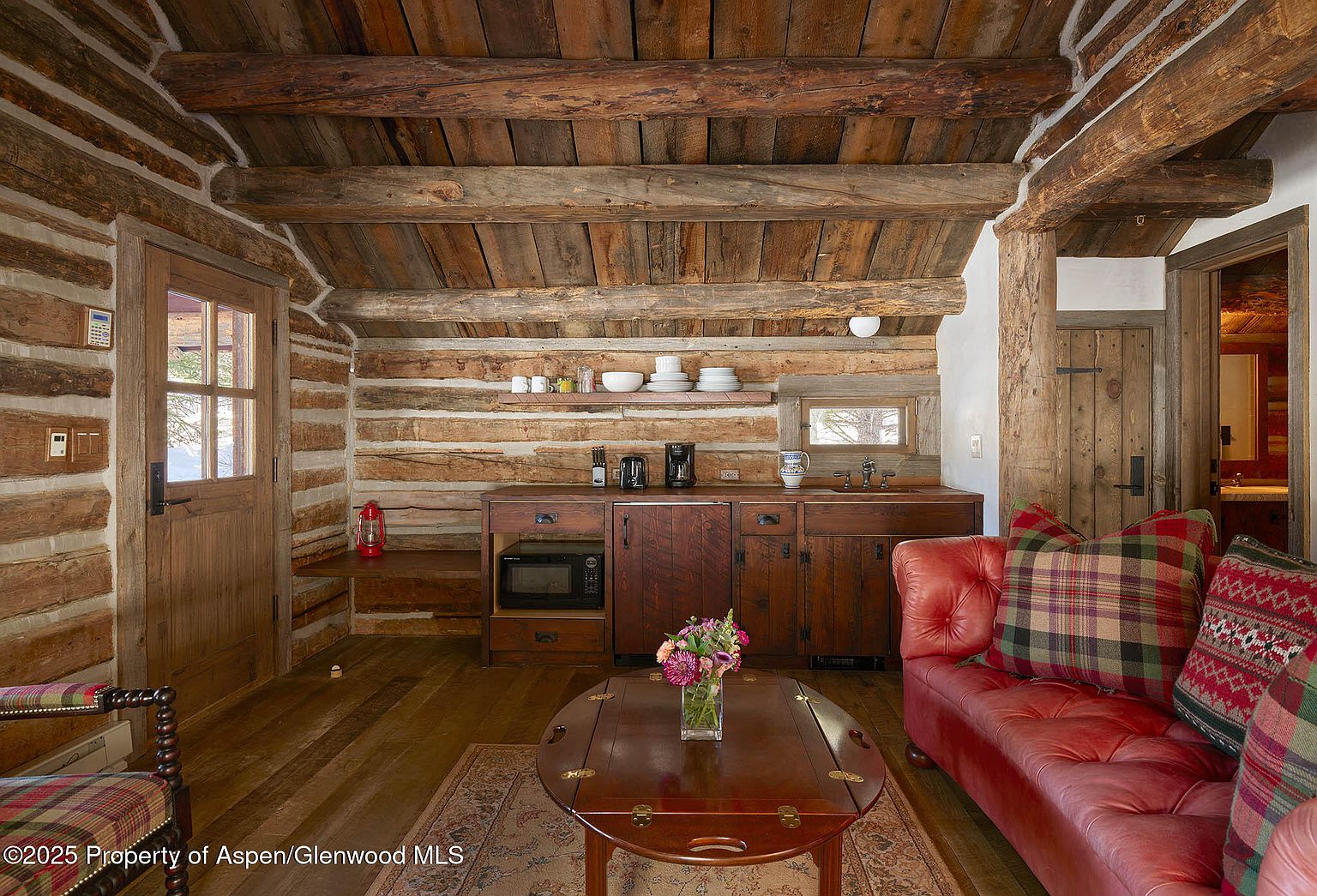 This interior shot showcases a rustic living room with log walls and a wooden ceiling, creating a cozy cabin atmosphere. A red leather sofa with plaid pillows sits in the foreground, complemented by a wooden coffee table with flowers. The room also features a kitchenette area with wooden cabinets and a small window, blending living and kitchen spaces seamlessly.