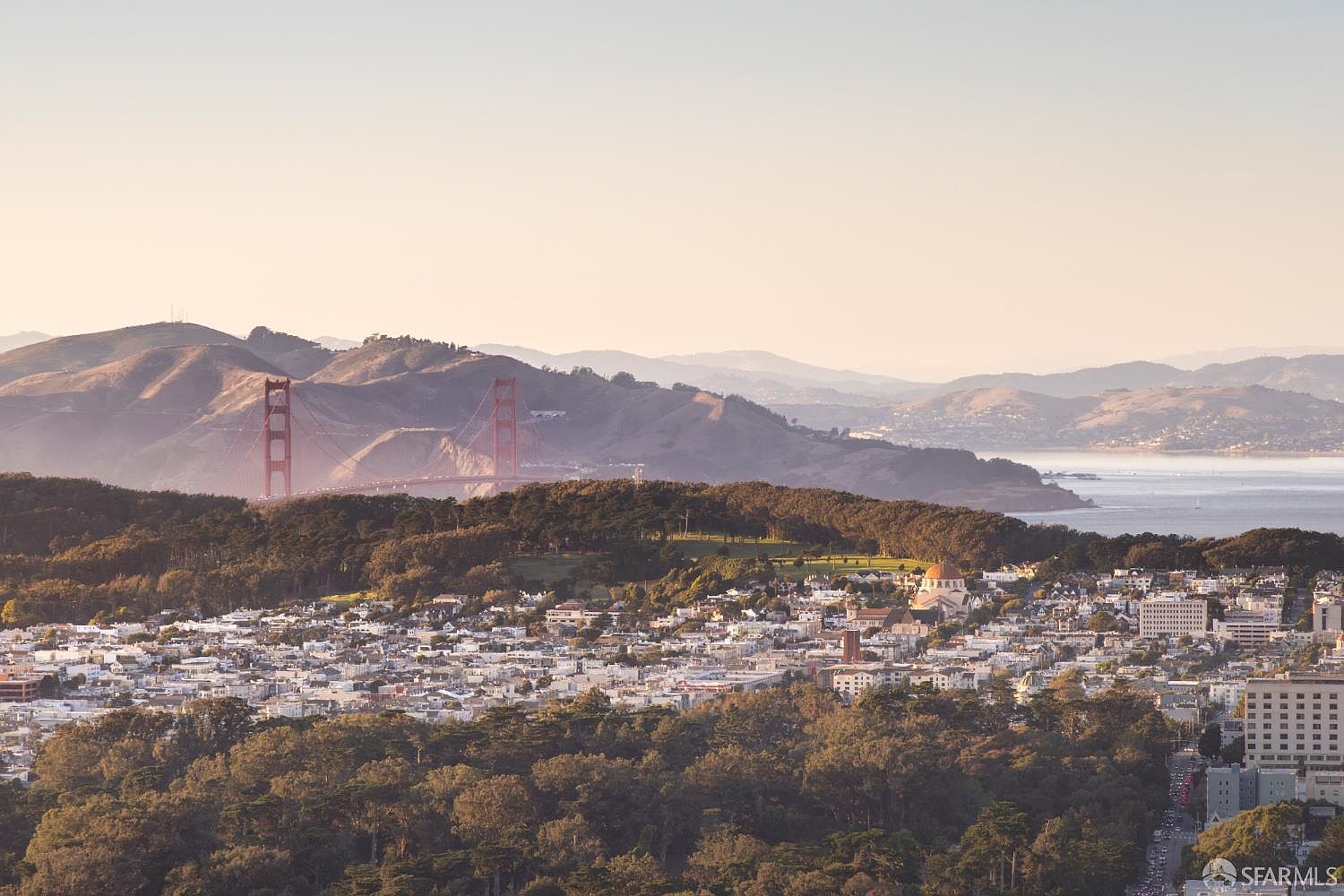 This aerial view captures a picturesque cityscape with a view of the Golden Gate Bridge in the background. The landscape is characterized by a mix of residential neighborhoods and lush green trees, with a glimpse of the water creating a serene atmosphere. The image showcases the density and natural beauty of the location, making it an appealing glimpse into the area's lifestyle.