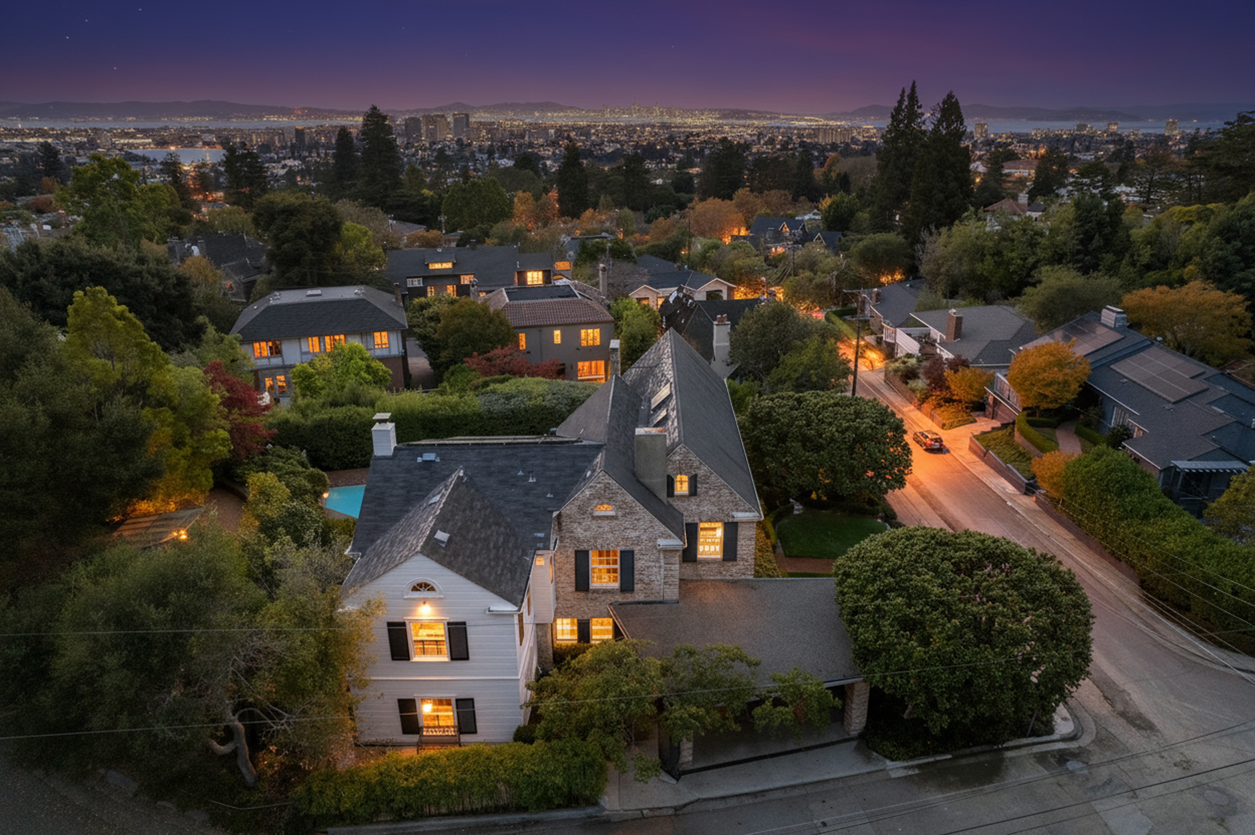 This aerial view showcases a stunning residential property at twilight, featuring meticulously maintained landscaping, elegant architectural details, and inviting illuminated windows. The setting includes a well-lit street and provides a glimpse of the city skyline in the background, adding to the home's appeal and desirability, making it an attractive real estate opportunity.