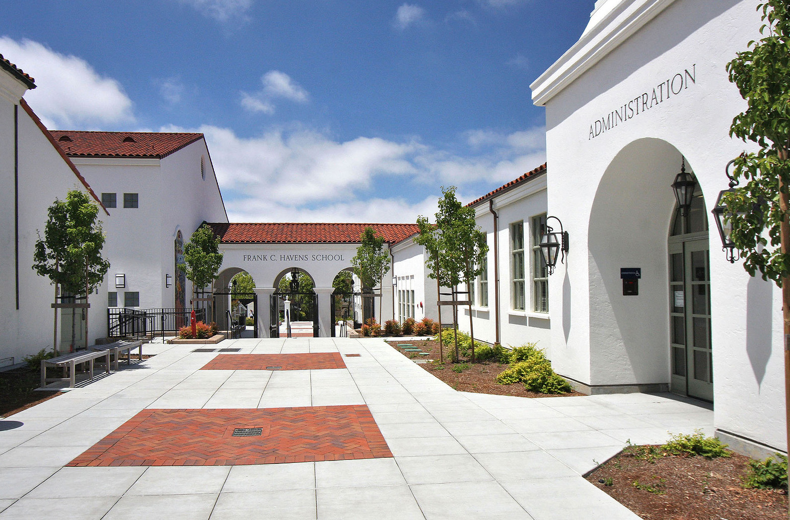 This is a front view of the Frank C. Havens School Administration building, presenting a well-maintained and inviting exterior. The architecture features a classic Spanish colonial style with white stucco walls, red tile roofing, and arched entrances. The landscaped courtyard with trees and brick accents complements the building's aesthetics, enhancing its curb appeal.