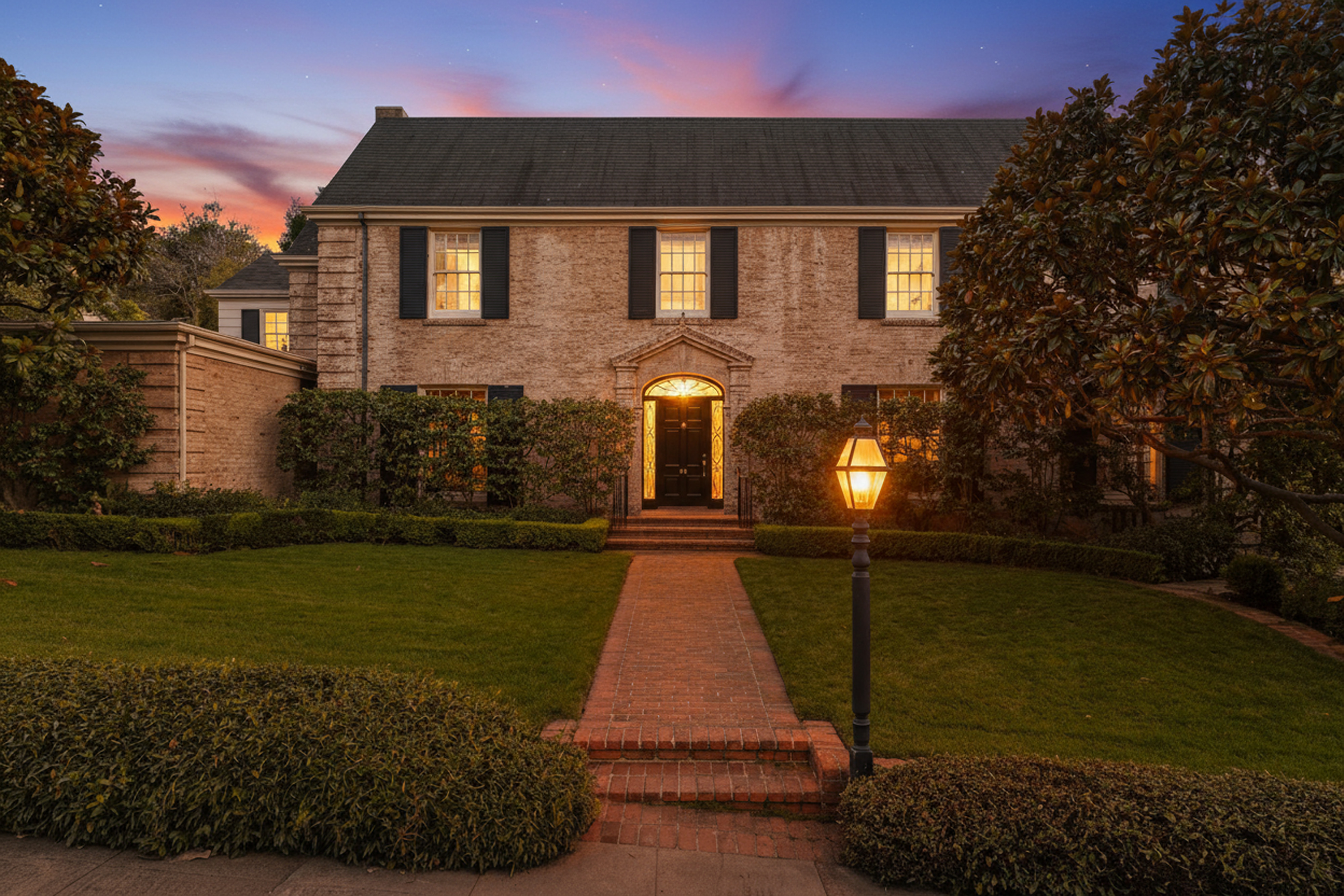 This is an inviting front view of a two-story brick home with elegant dark shutters. A well-manicured lawn and mature landscaping frame the property, while a brick pathway leads to the dark, arched entryway. The warm lighting from the windows and a lantern creates a welcoming ambiance.