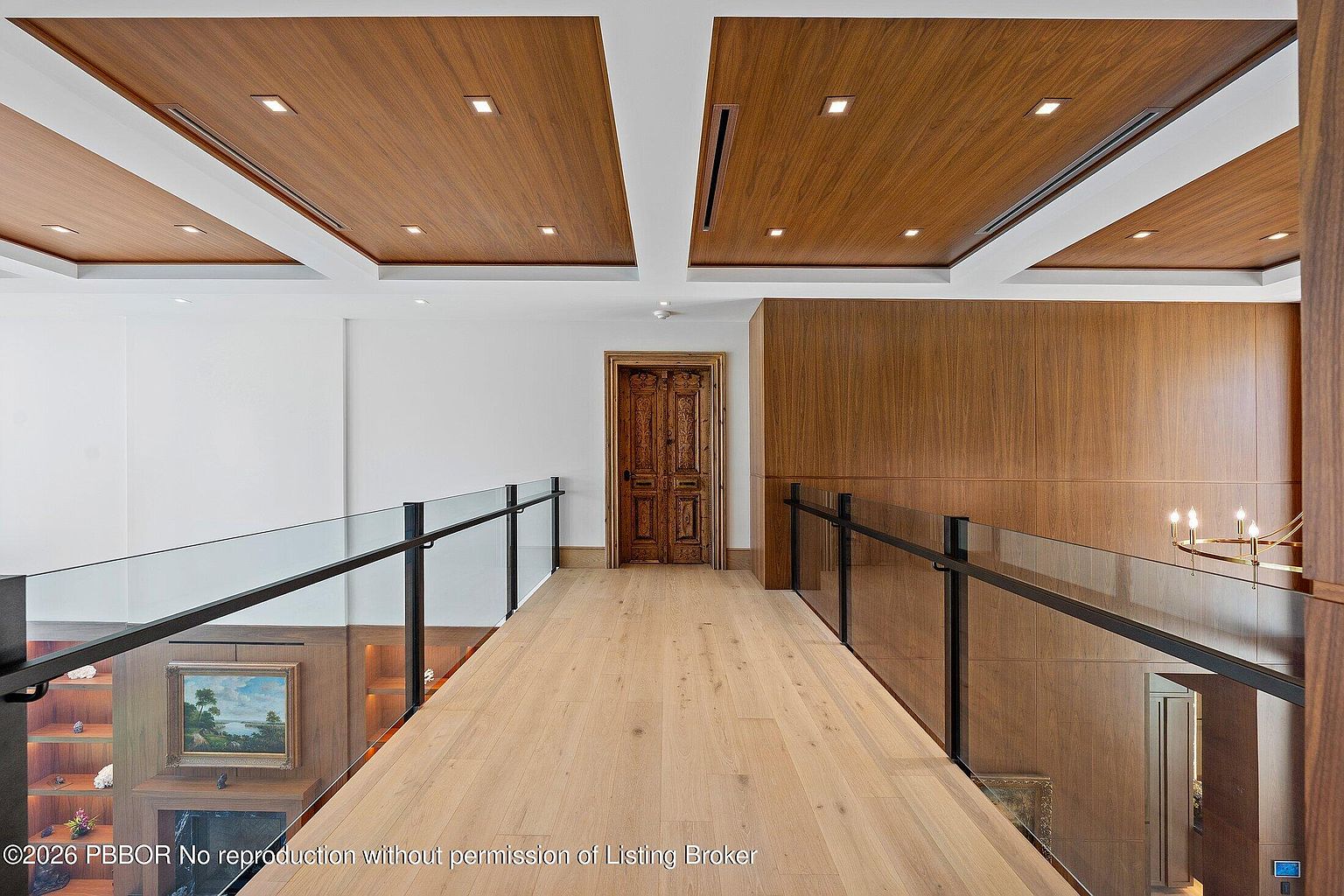 This interior view captures a sophisticated bridge-style hallway overlooking a lower level, defined by a glass railing and light wood flooring. The ceiling features recessed lighting set within wood-paneled coffers, while a decorative, ornate wooden door serves as a focal point at the end of the walkway. The perspective is elevated and symmetrical, emphasizing the clean lines and high-end architectural details of the home.