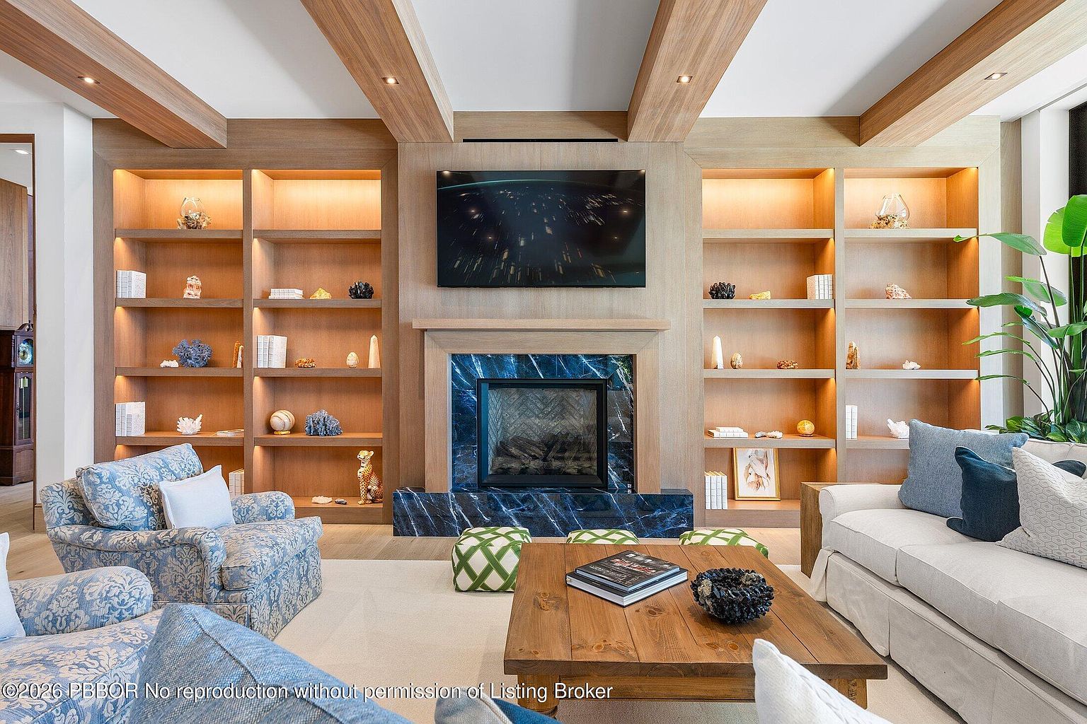 This elegant living room features a central fireplace with a striking blue stone surround, flanked by extensive light wood built-in shelving units. The space is furnished with a plush sofa, patterned armchairs, and a rustic wooden coffee table, creating a sophisticated yet comfortable atmosphere. The room is accented by exposed wooden ceiling beams and decorative objects displayed on the shelves, offering a warm and inviting aesthetic.