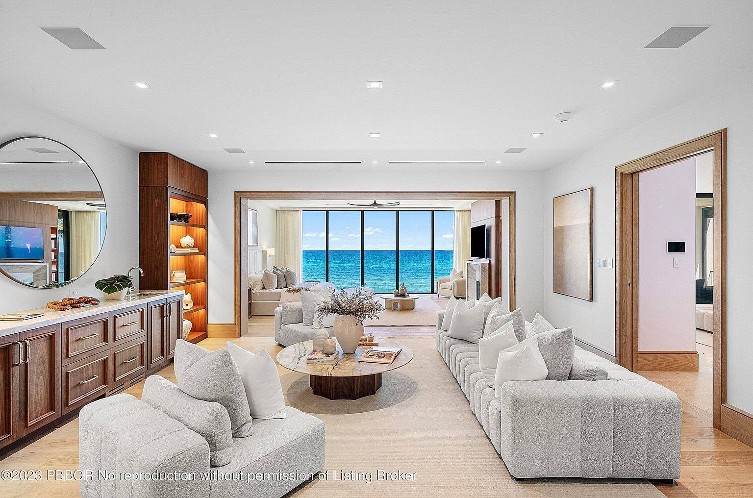 This sophisticated living area features a neutral color palette with plush, textured seating arranged around a circular stone-topped coffee table. A built-in wood shelving unit and a wet bar with a large circular mirror anchor the side of the room, while an open transition leads to a secondary living space with expansive glass walls overlooking the ocean. The design emphasizes a seamless flow between indoor comfort and the scenic coastal view beyond.